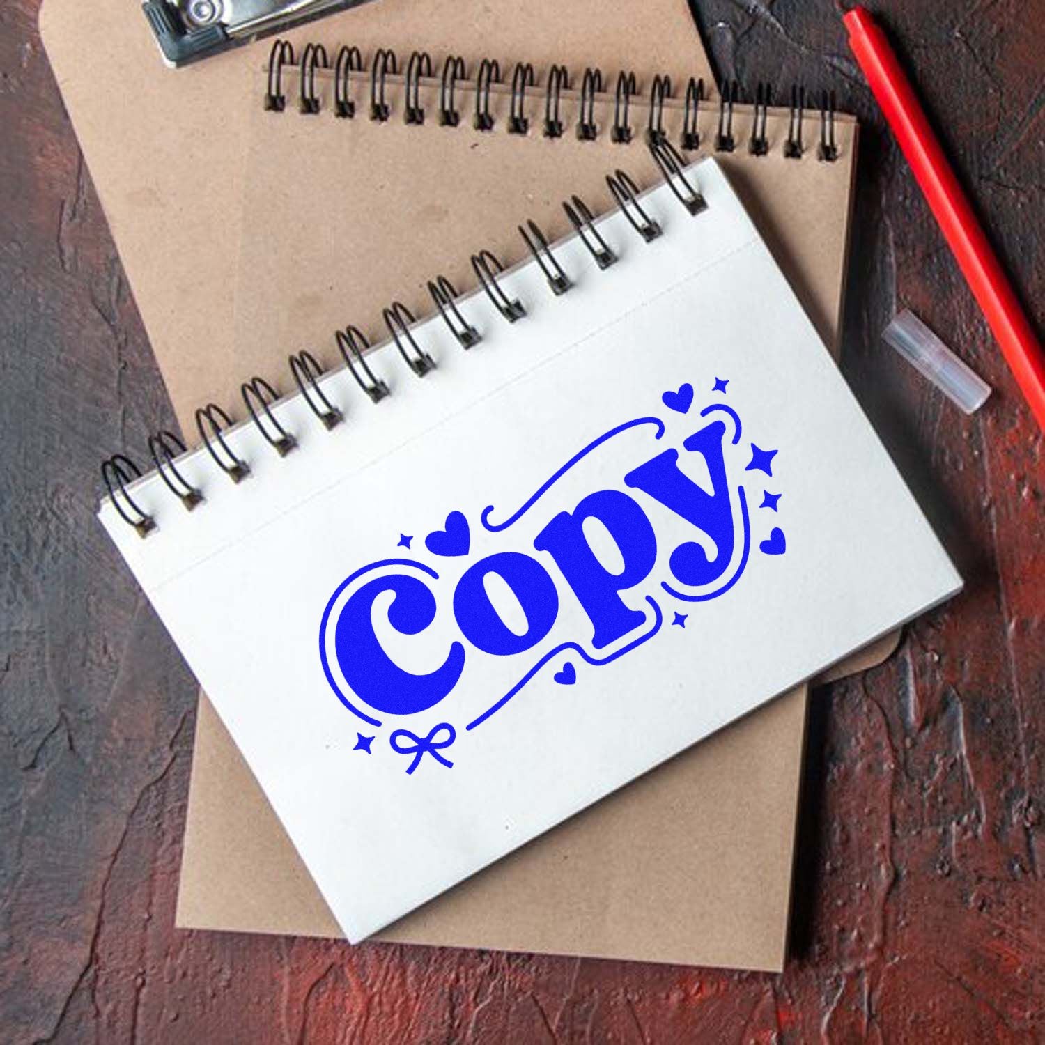 Blue COPY stamp seal on a white spiral notebook, with hearts and stars, over a kraft notebook on a dark wood desk.