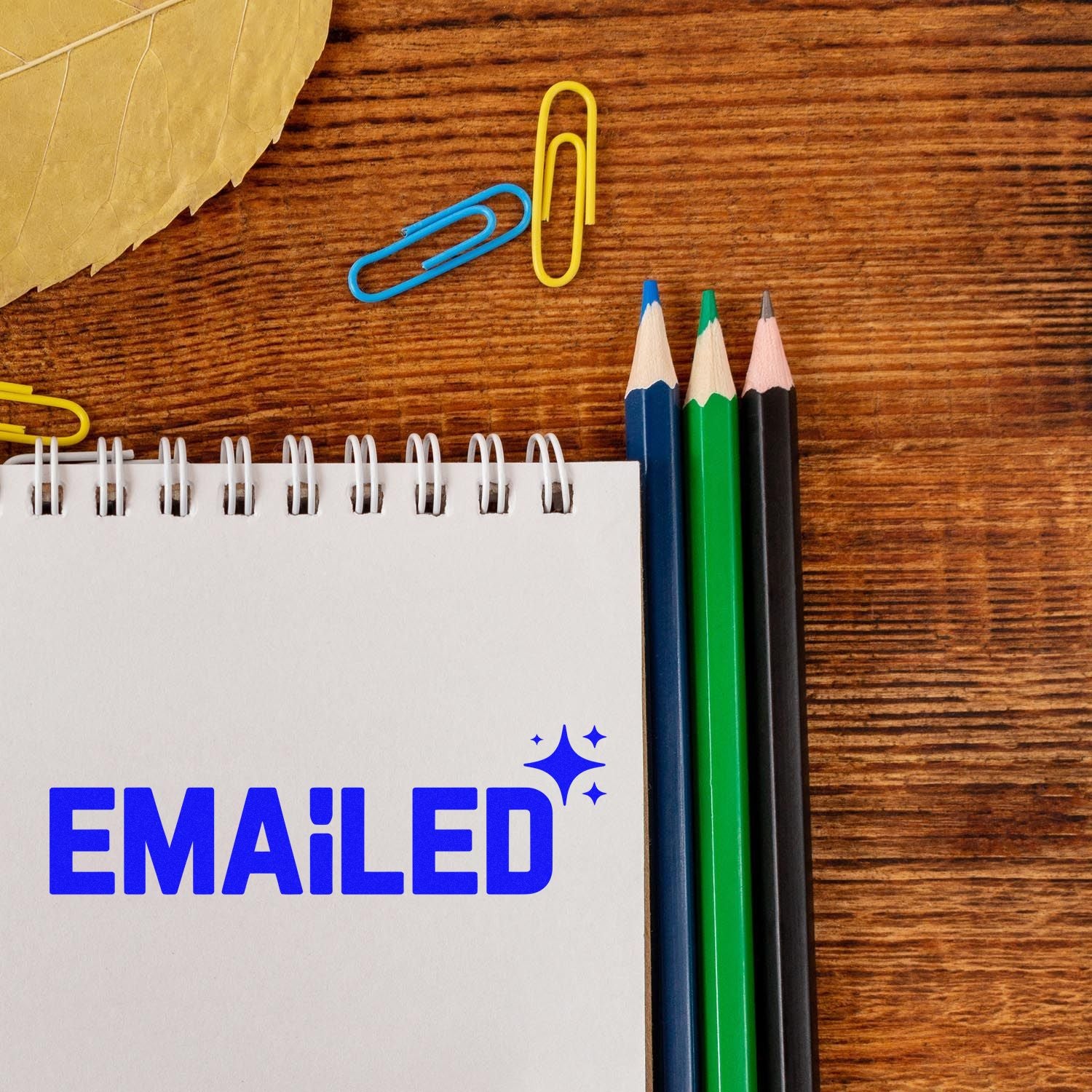 Blue EMAILED rubber stamp and seal mark on notepad, with colored pencils and paperclips on a wood desk.