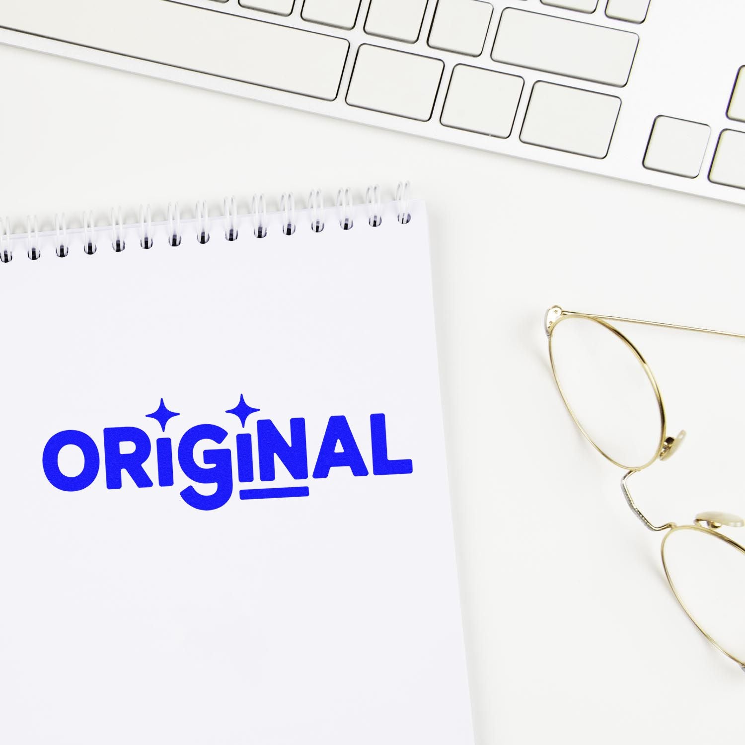 Blue ORIGINAL stamp seal on a white spiral notepad beside gold glasses and a white keyboard