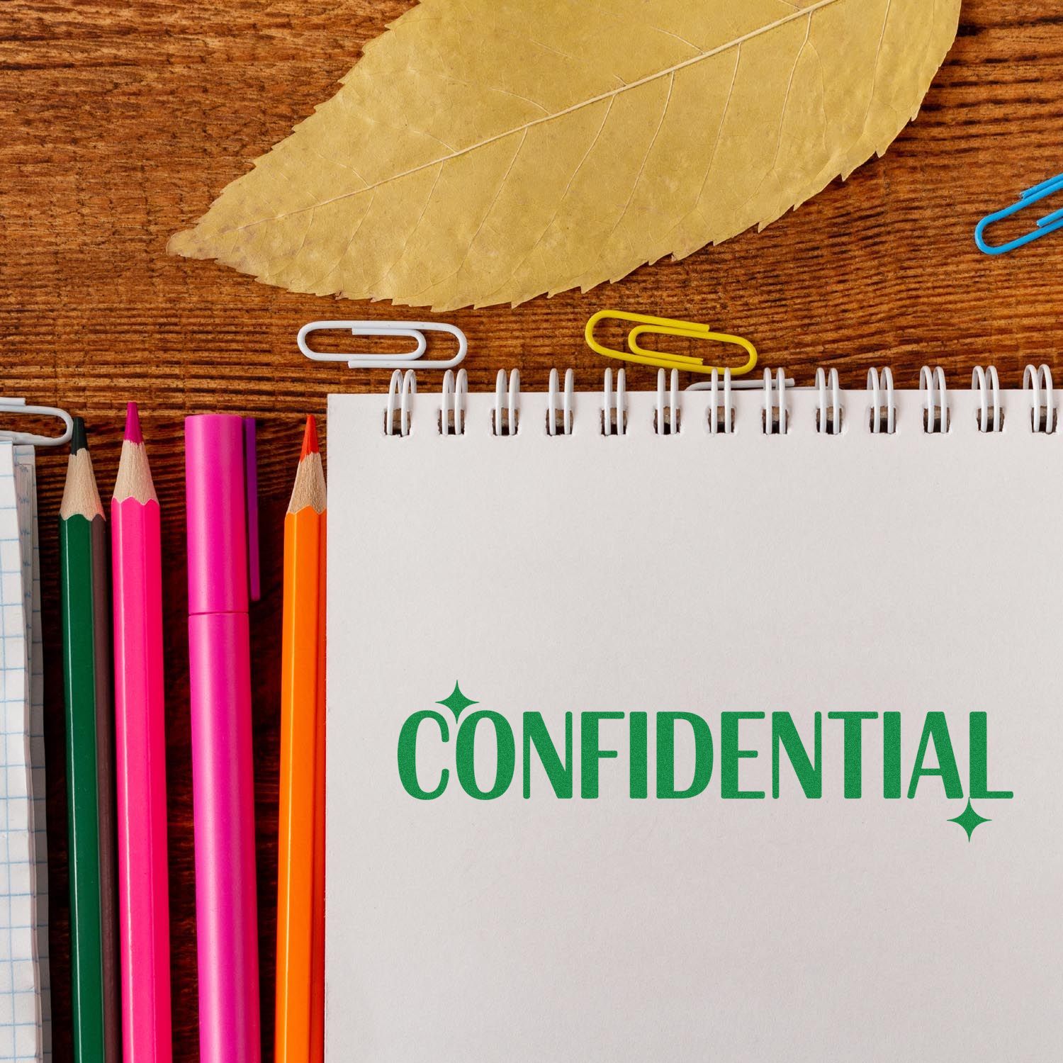 Green confidential stamp on a white spiral notebook, with colorful pencils, paper clips and a dried leaf on wood.