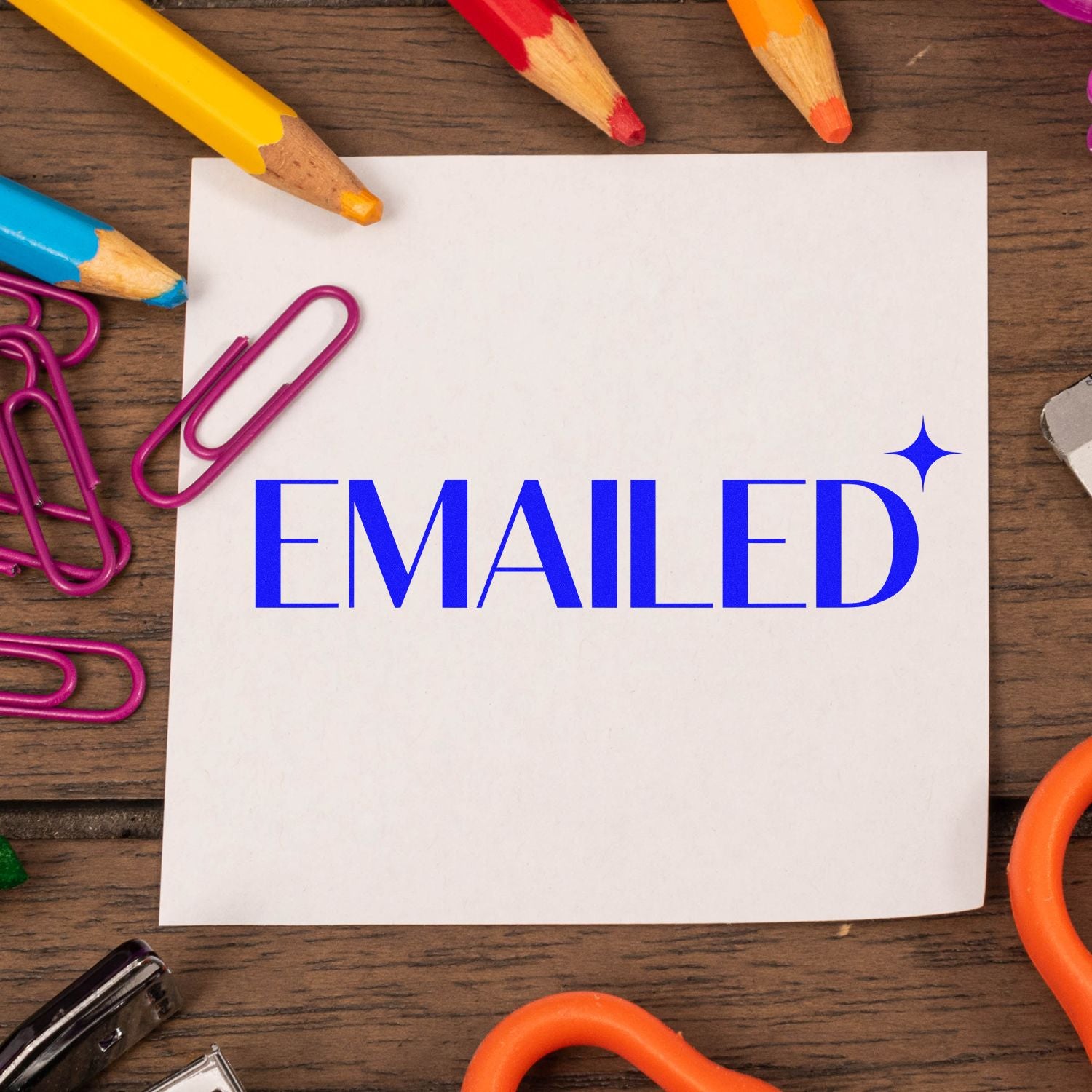 Blue EMAILED seal stamp on white paper, surrounded by colorful pencils and magenta paper clips on a wooden desk.