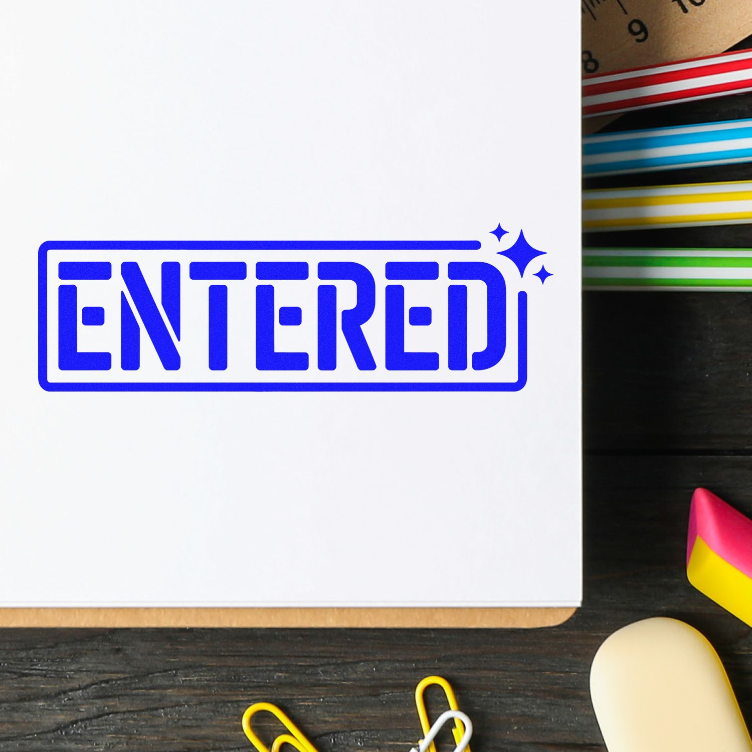 Blue ENTERED approval stamp with sparkles on white paper, beside colorful seal markers and stationery.