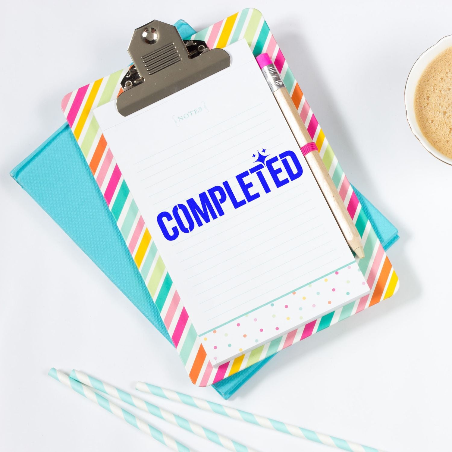 Colorful striped clipboard with blue COMPLETED stamp, lined notepad, pencil, and turquoise notebook beside coffee.