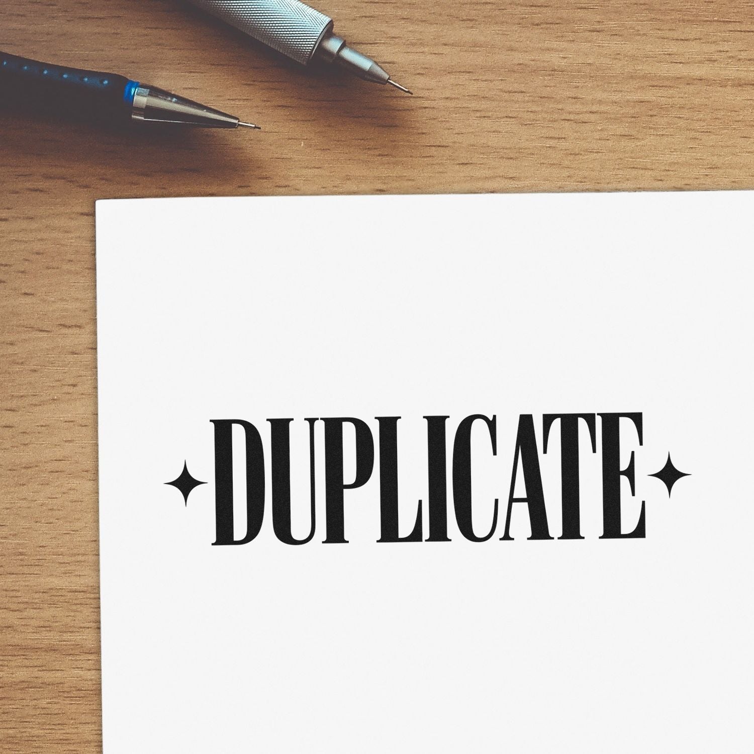 Black DUPLICATE stamp with star seal marks on white paper, beside two pens on a light wood desk.