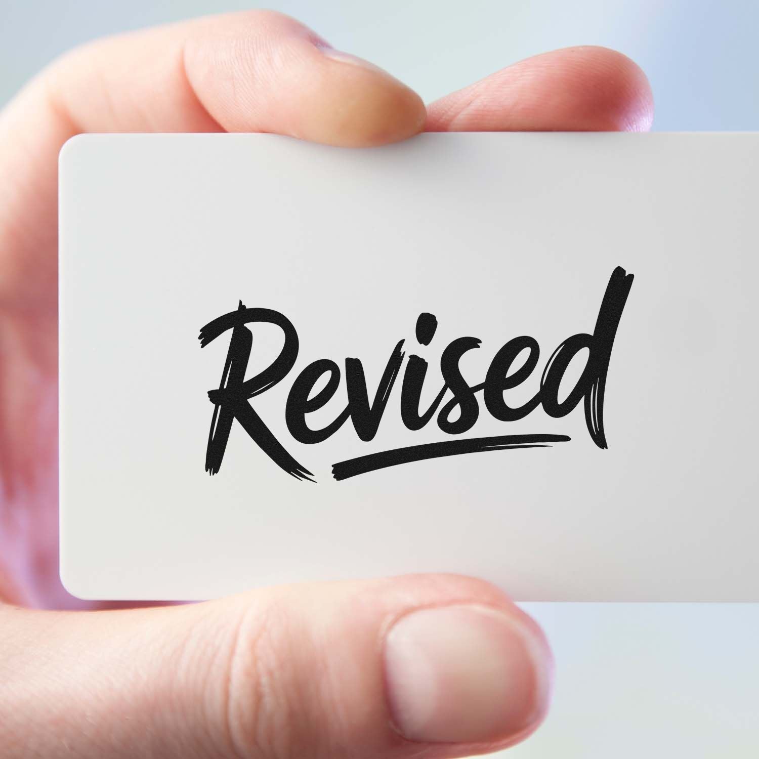 Hand holding a white stamp and seal card with black Revised text on a clean light background.