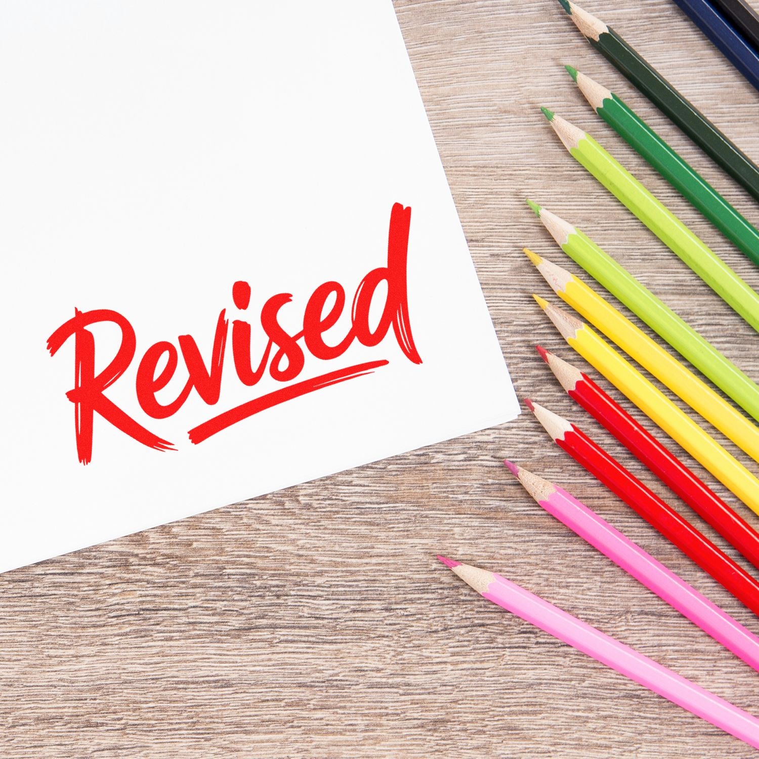 Red revised rubber stamp seal on white paper beside colorful pencils on a wood desk