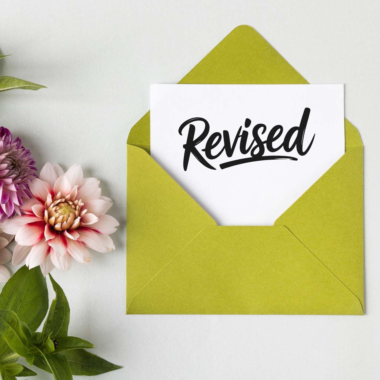 Green envelope with white card stamped Revised in black script, accented by pink and purple flowers.