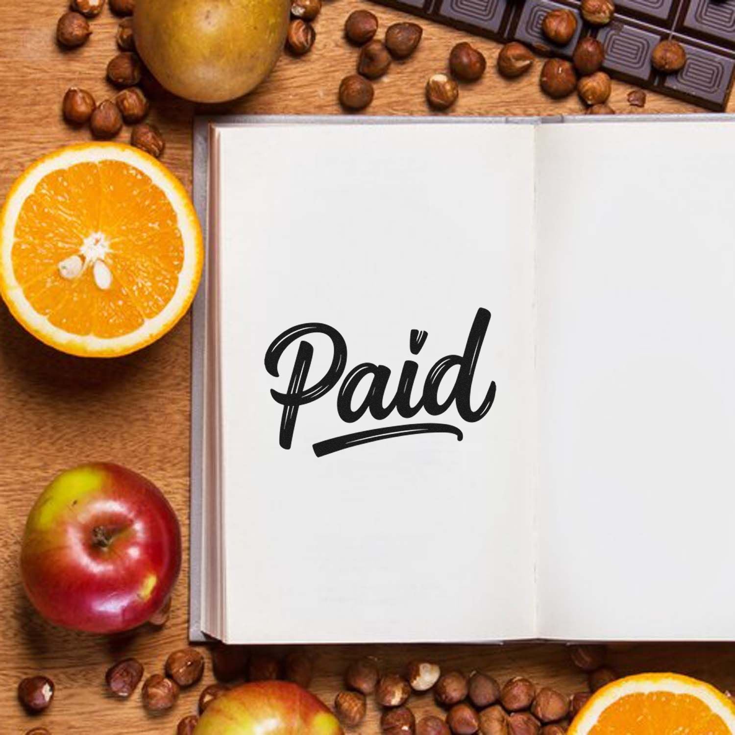 Black Paid stamp seal on open white notebook, with oranges, apples, and coffee beans on a wooden table.