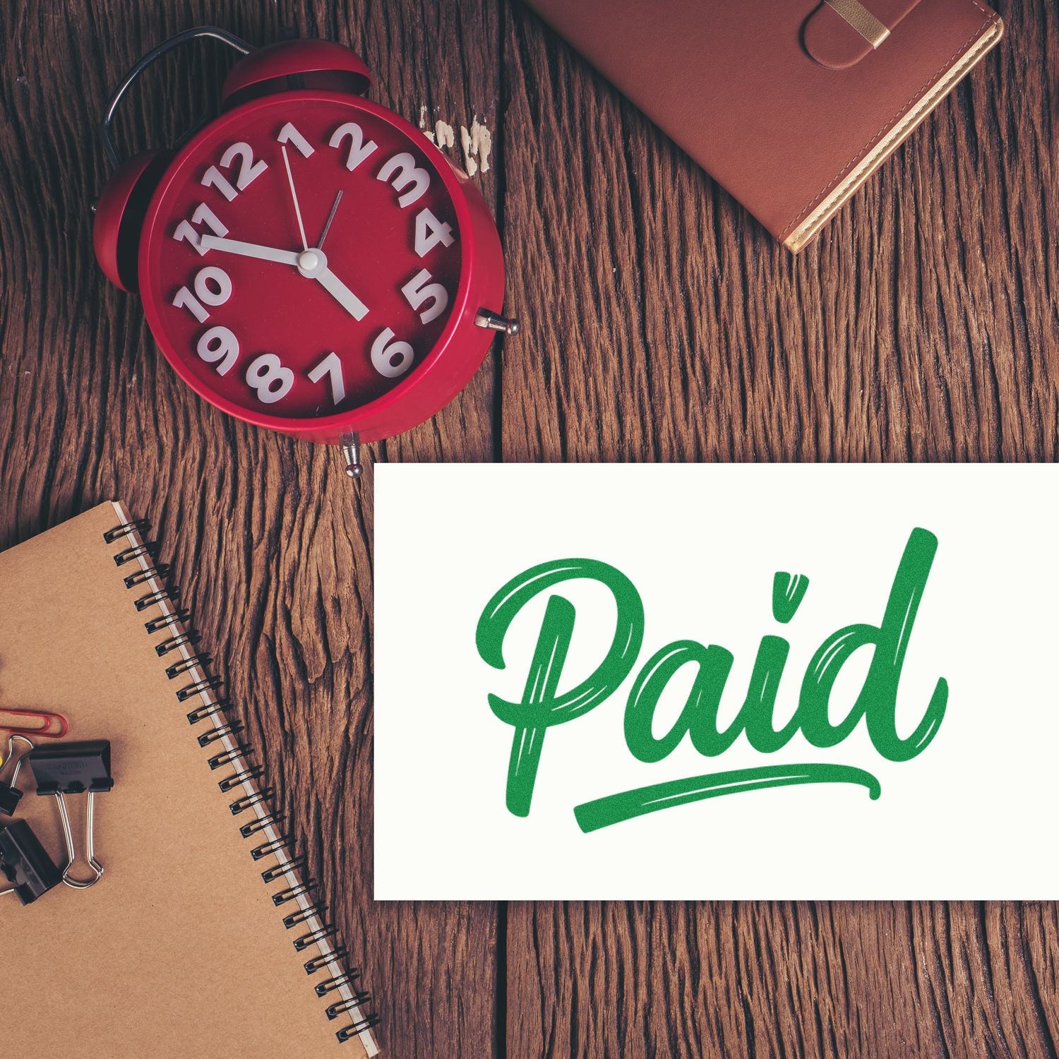 Green Paid stamp seal on white card with red alarm clock and brown notebook on a wood desk.