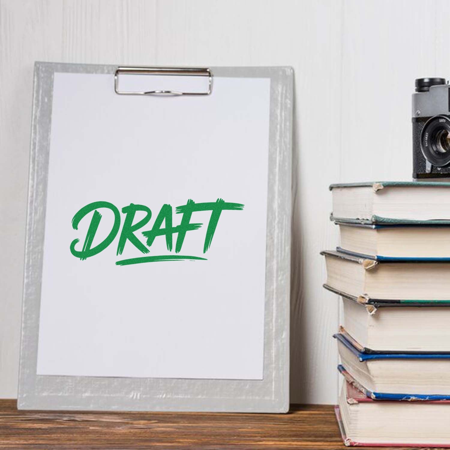 Clipboard with blank paper and green DRAFT stamp impression, beside stacked books and a vintage camera.