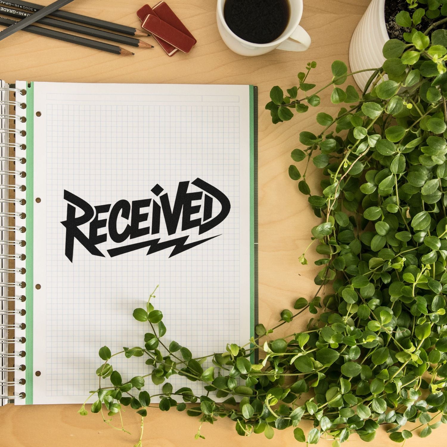 Black RECEIVED stamp seal on graph-paper notebook beside green plant, pencils, and coffee on a wood desk.