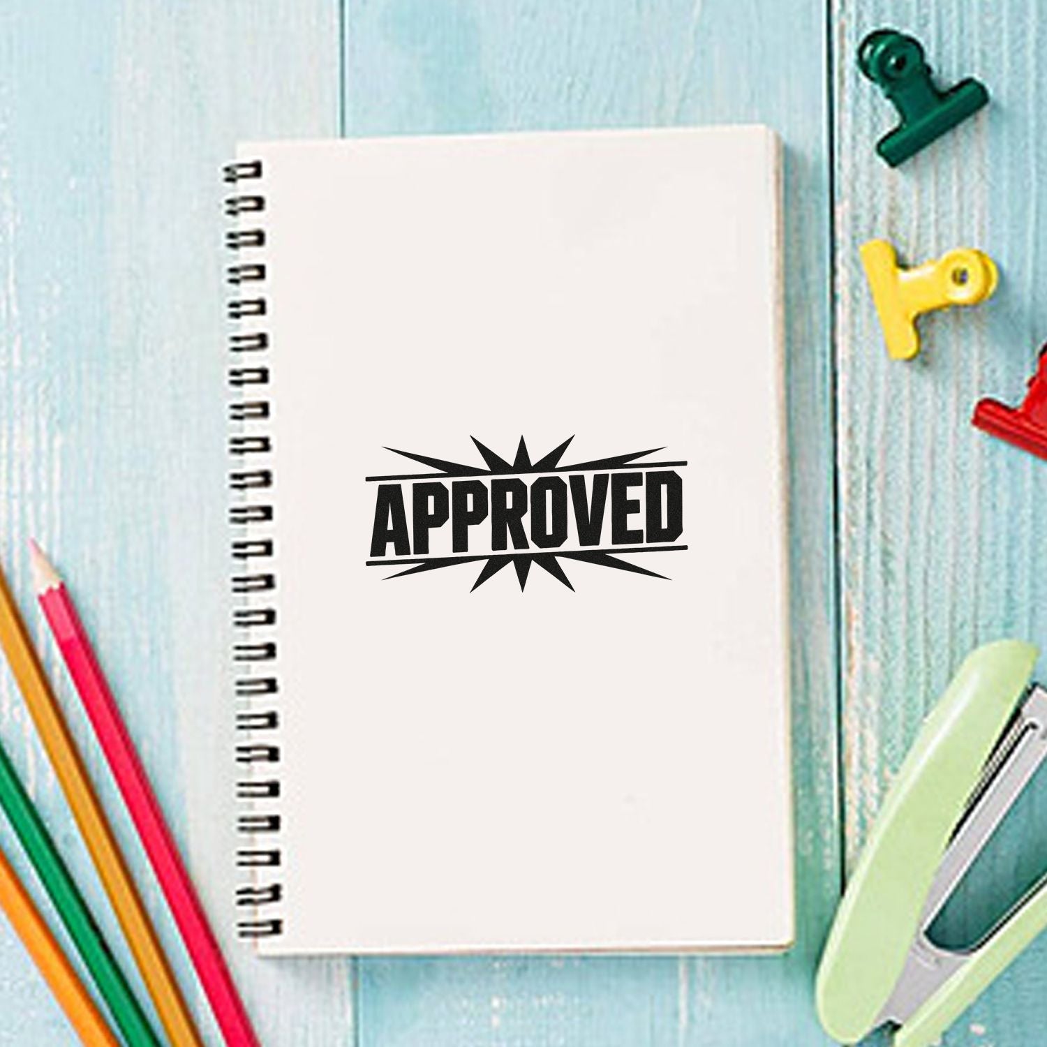 Black APPROVED stamp seal on a white spiral notebook, surrounded by colorful pencils and binder clips on a blue desk.