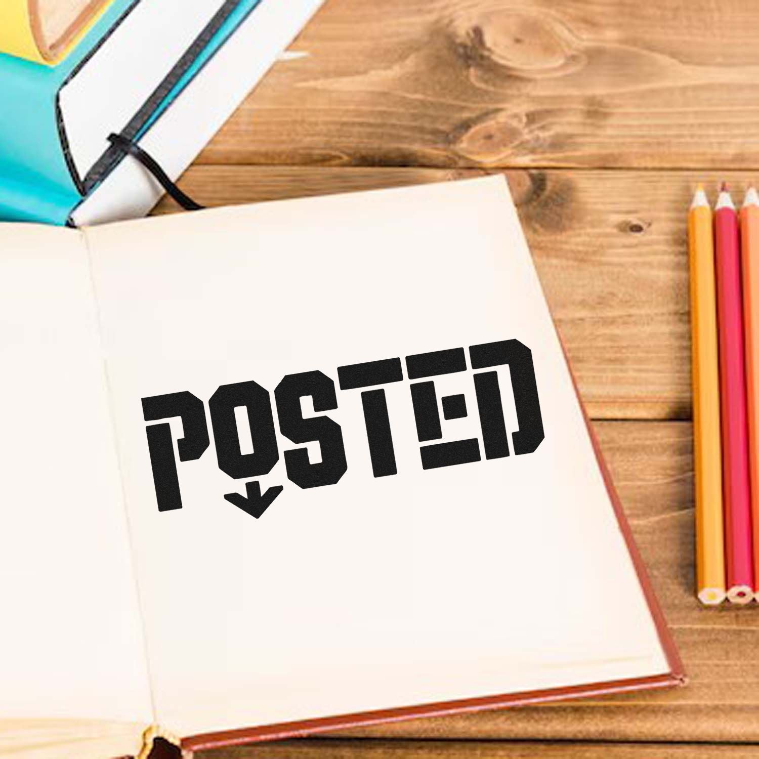Black POSTED rubber stamp seal on an open white notebook, with colorful pencils and teal notebooks on a wooden desk.