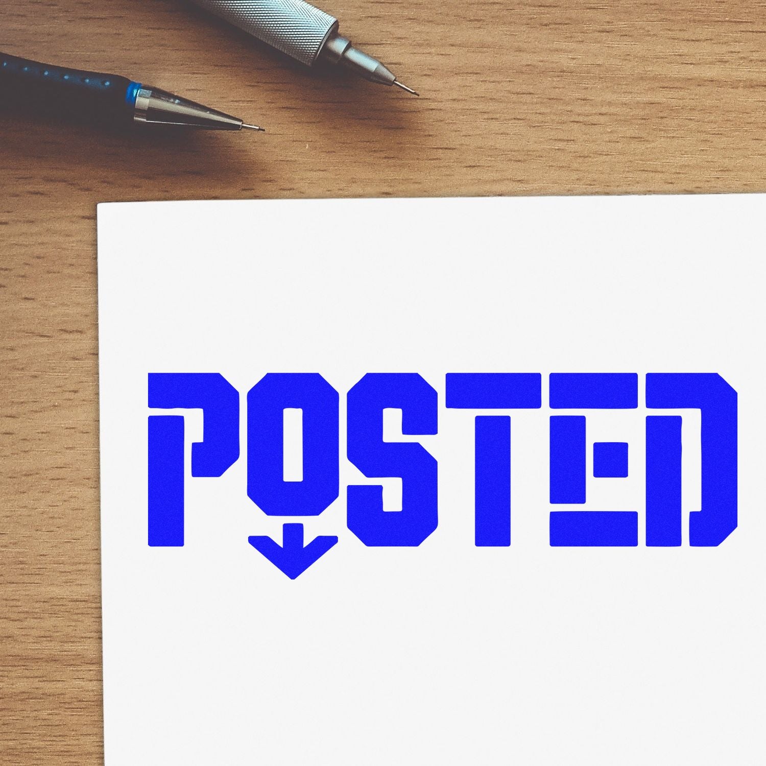 Blue POSTED stamp seal on white paper, with two pens on a wooden desk.