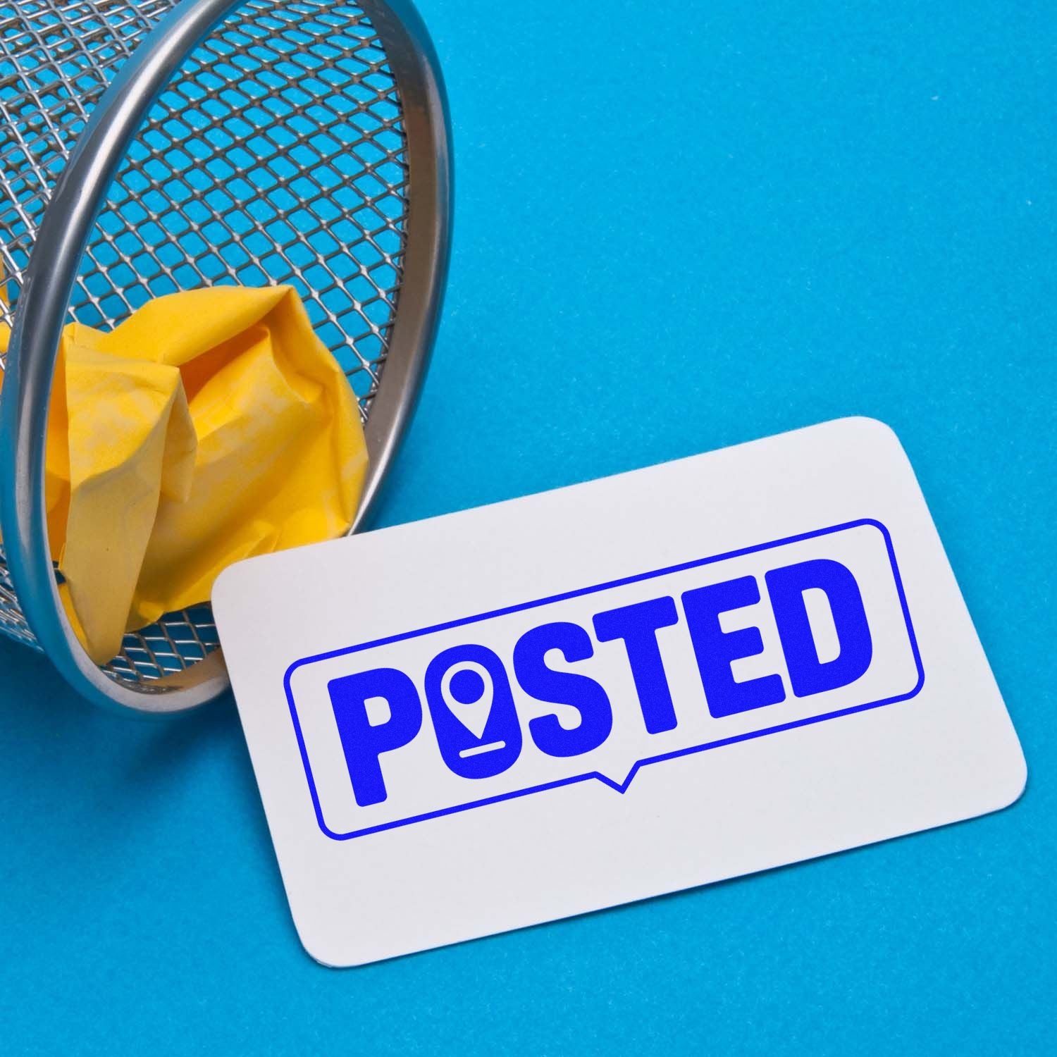 Blue POSTED stamp and seal card beside a silver mesh basket with crumpled yellow paper on a blue background.