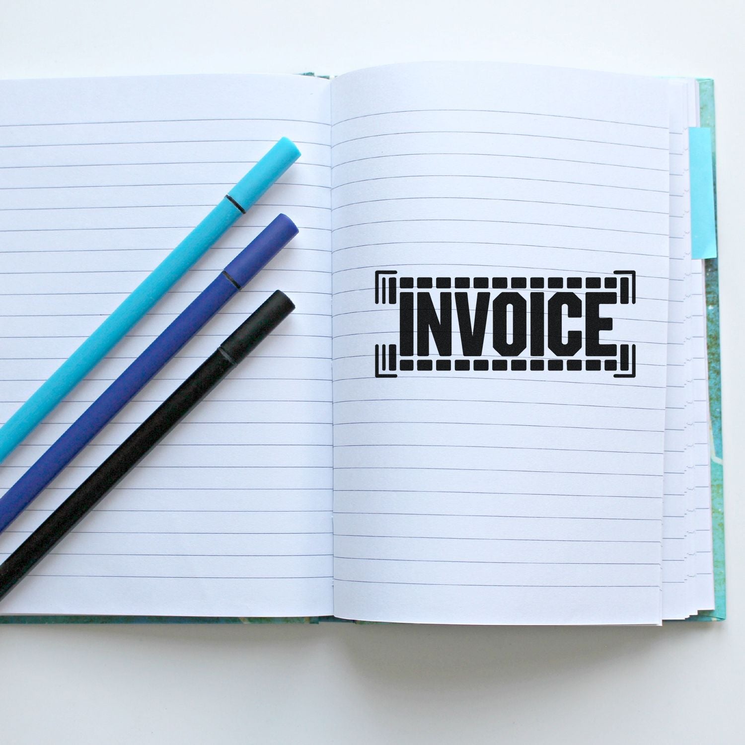 Black INVOICE stamp seal on open lined notebook, with turquoise, blue, and black pens.