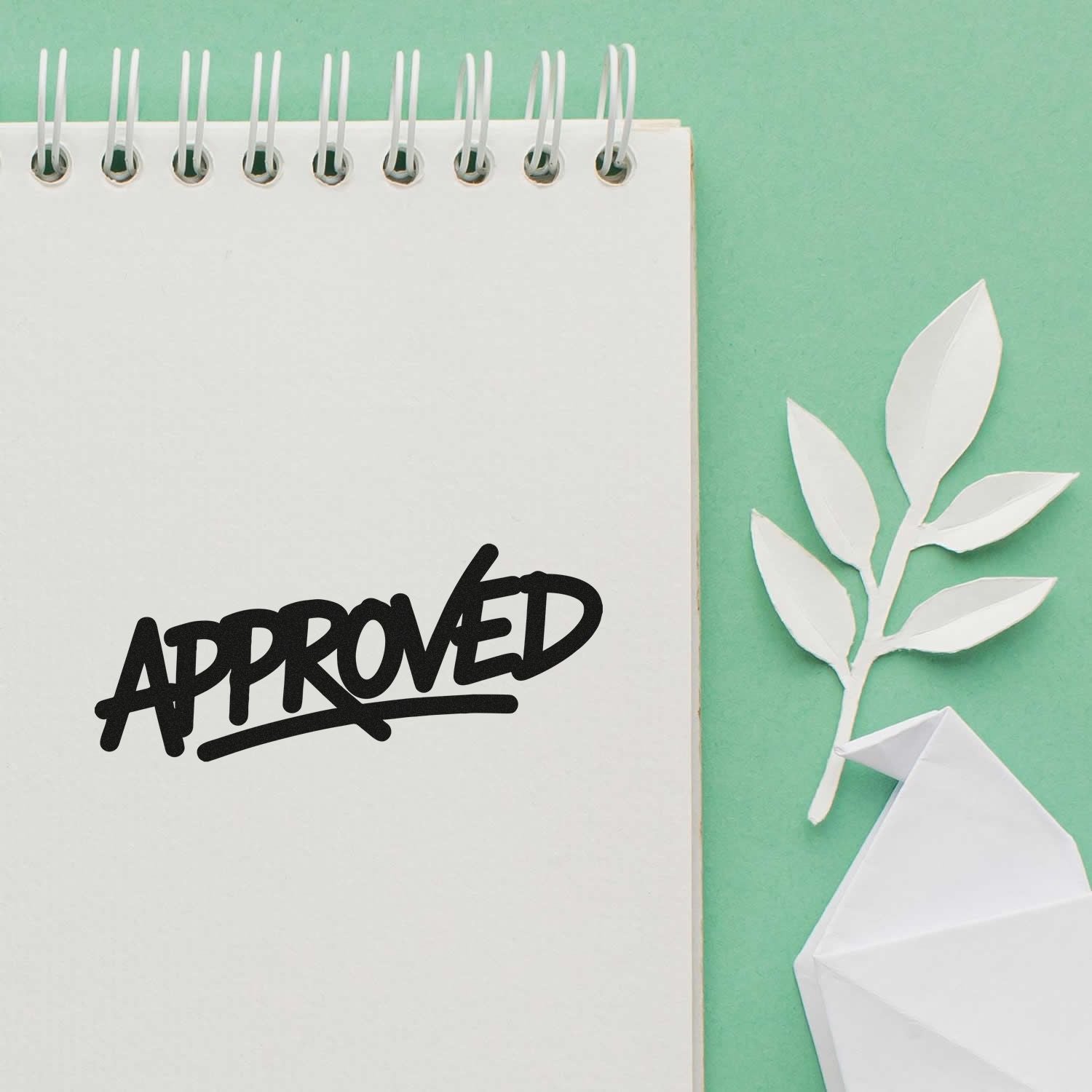 Black APPROVED stamp on white spiral notepad with white paper leaves and folded seal on mint green background