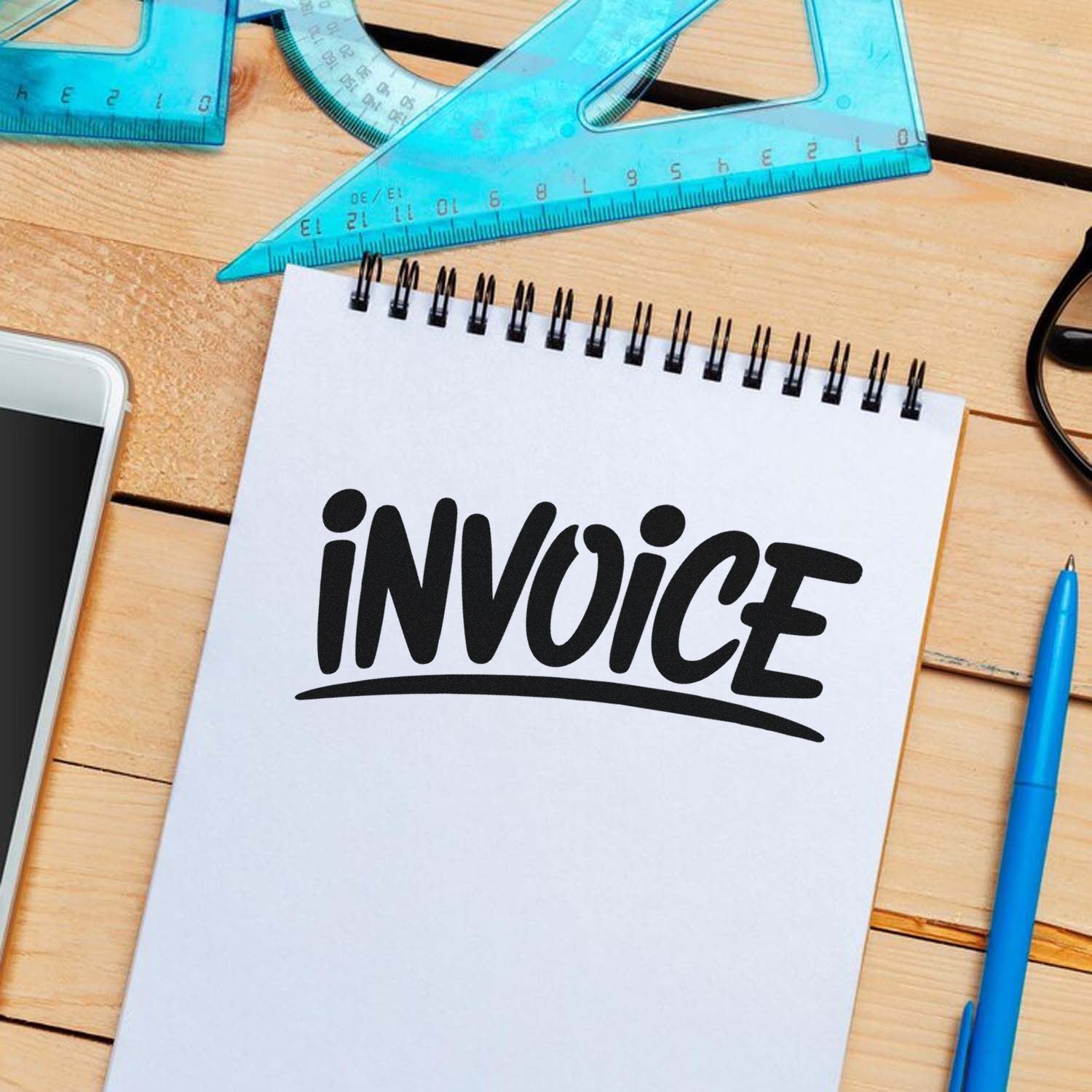 Black INVOICE stamp and seal design on white notepad with blue rulers, pen and phone on a wooden desk.
