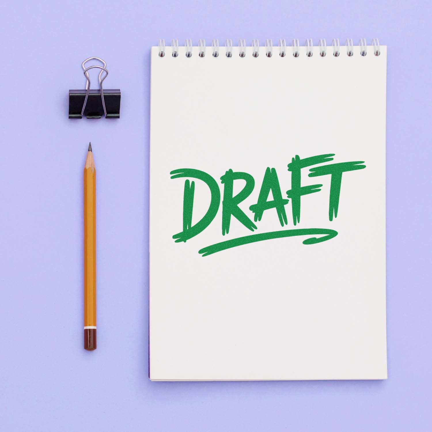 Green draft stamp on a white spiral notebook, with a pencil and black binder clip on a lavender background.