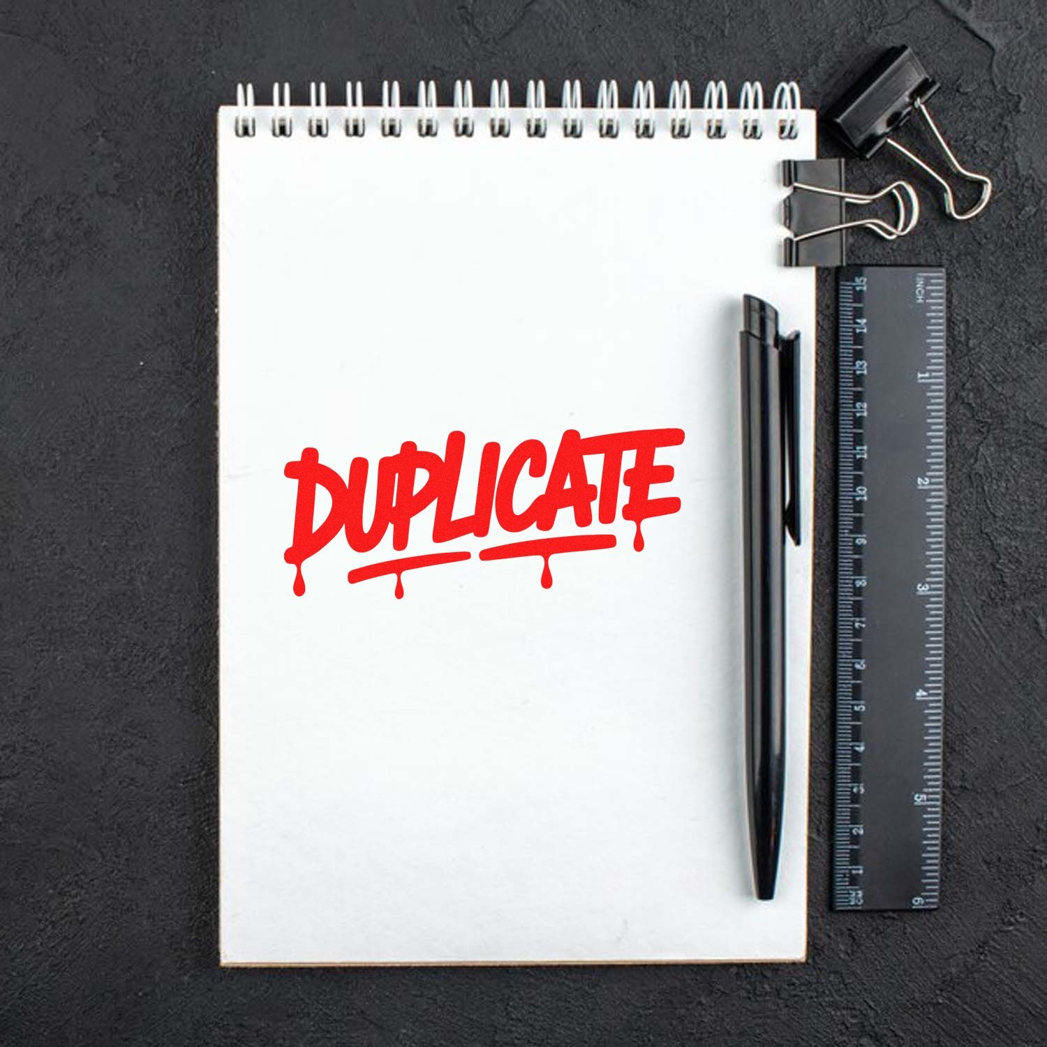 Red DUPLICATE stamp on white spiral notepad with black pen, ruler, and binder clips, showcasing stamp and seal supplies.