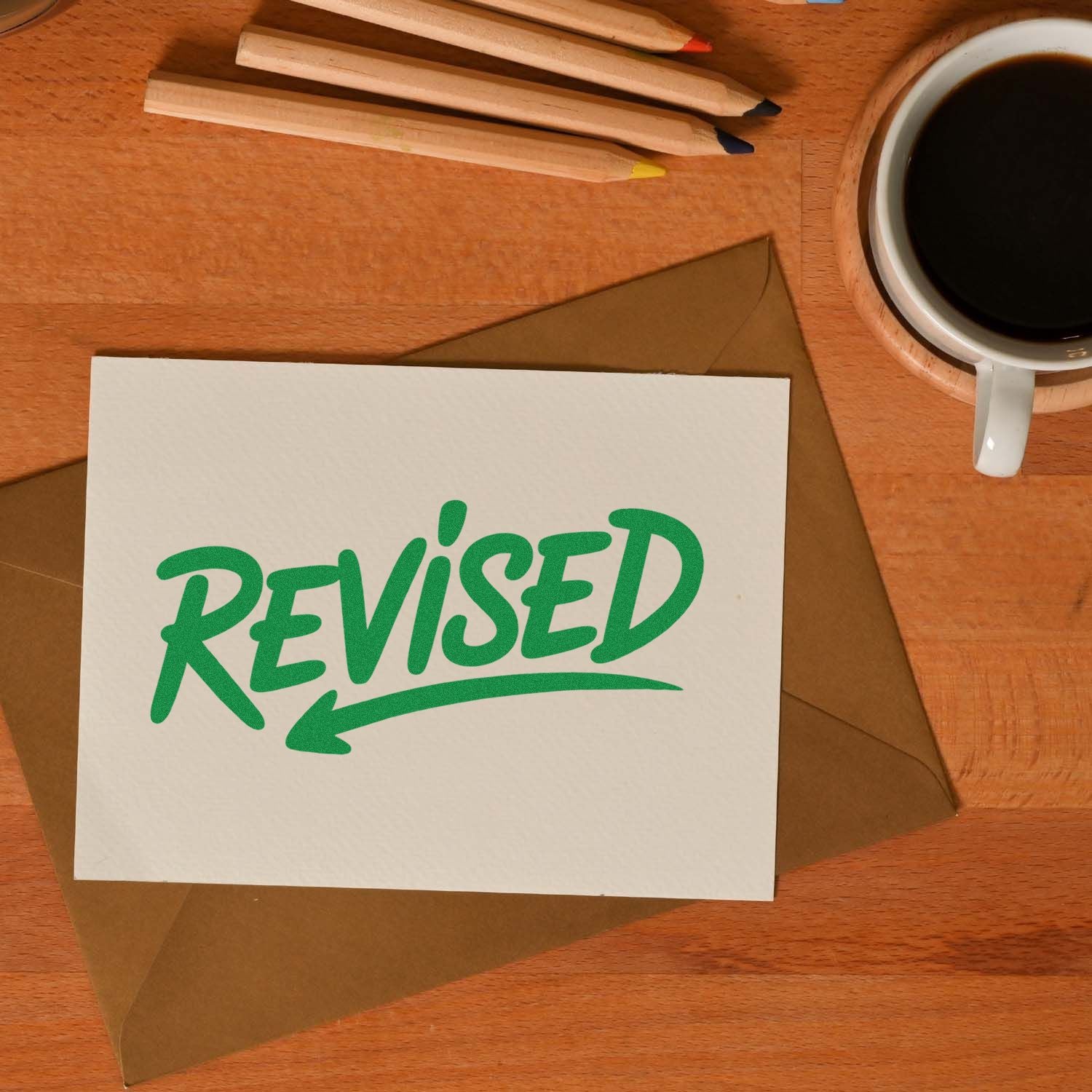 Green REVISED rubber stamp impression on white card over brown envelope, with pencils and coffee on a wooden desk.