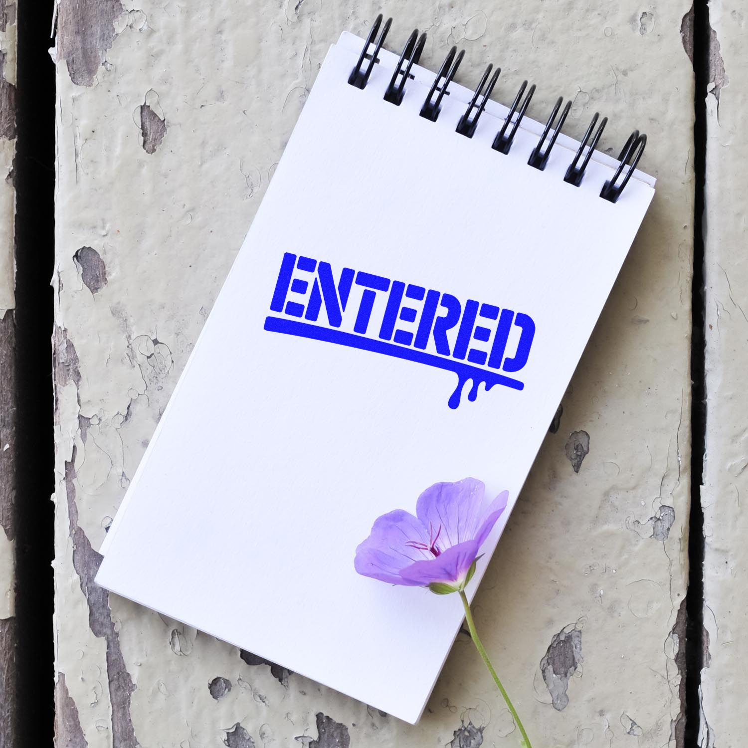 White spiral notepad with blue ENTERED stamp seal graphic and a purple flower on weathered gray wood.