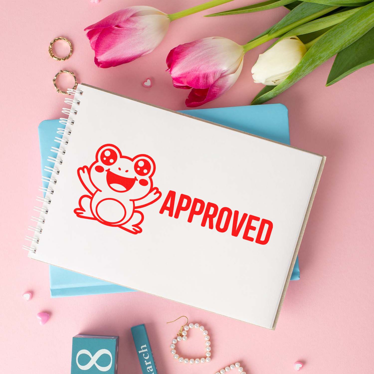 Red approved frog seal stamp on a white spiral notebook, with pink tulips and blue folders on a soft pink background.