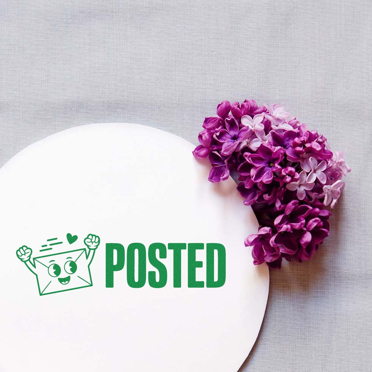 White round card with green POSTED stamp seal and cartoon envelope, beside purple lilac flowers on gray fabric.