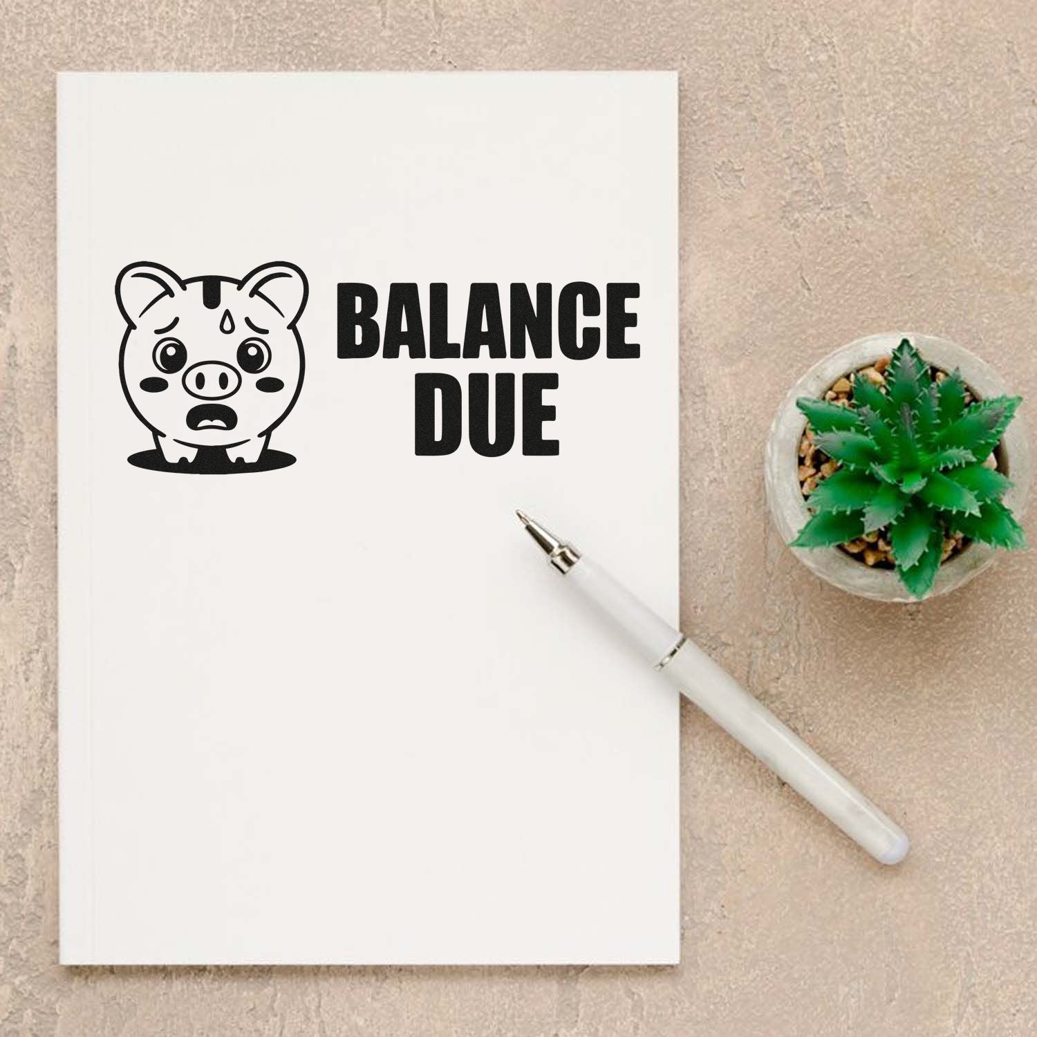 Black balance due stamp seal with worried pig icon on white paper, beside a white pen and green succulent.