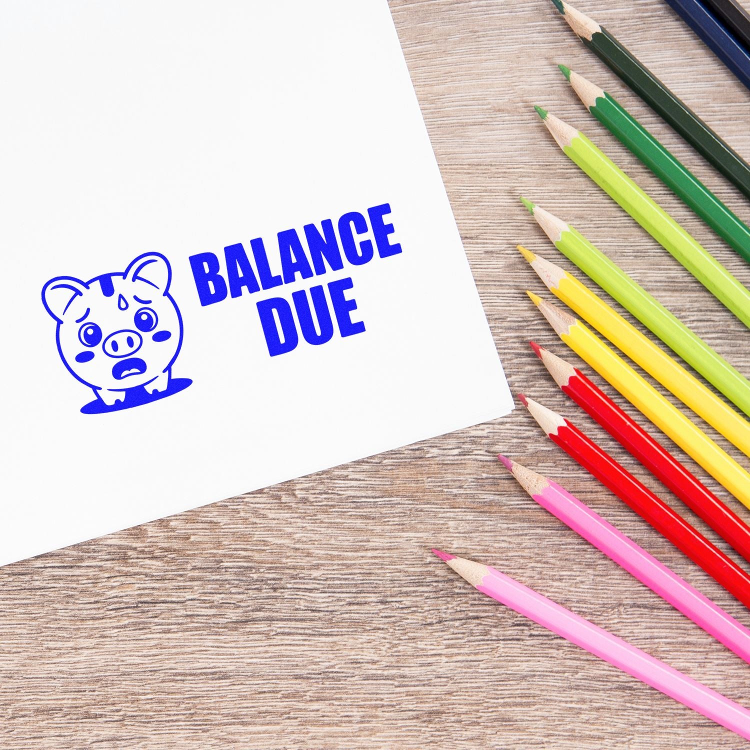 Blue balance due stamp with pig seal on white paper beside colorful pencils on wood desk.