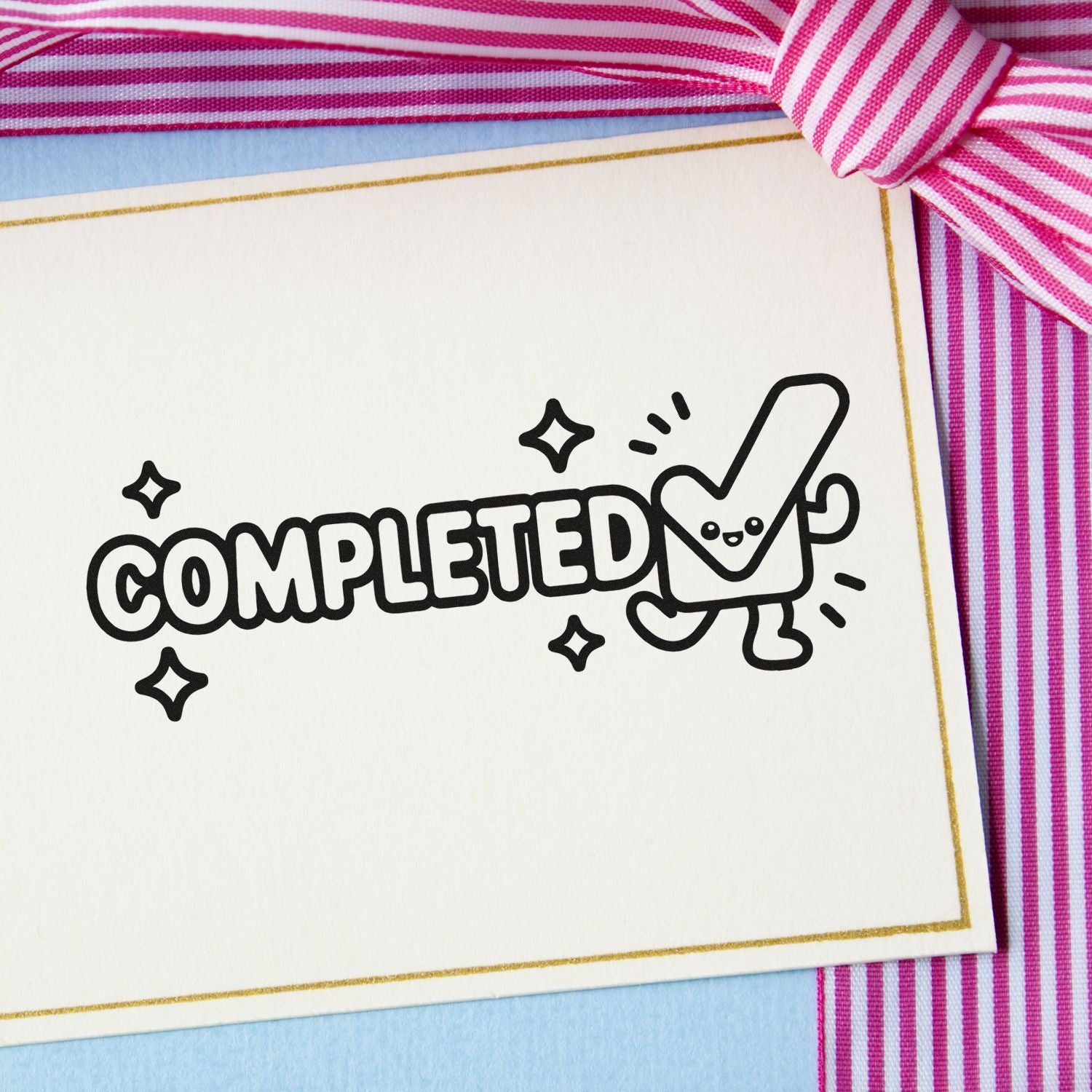Black COMPLETED stamp and seal on white card with gold border, wrapped in pink striped ribbon on blue background.