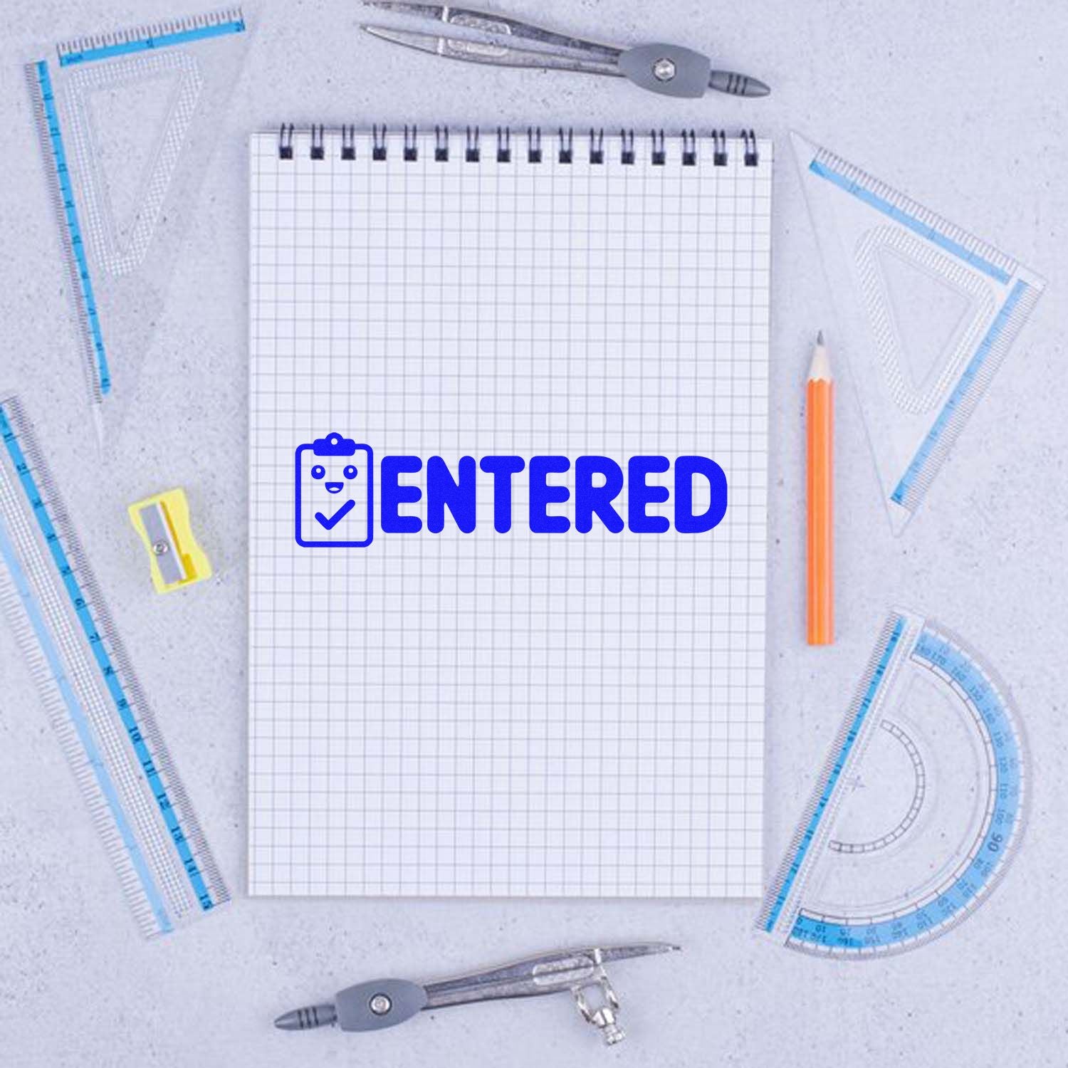 Blue ENTERED stamp seal on graph notebook, surrounded by clear rulers, compasses, and an orange pencil.