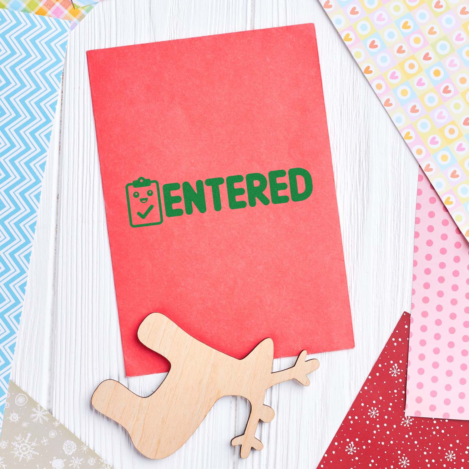 Red cardstock with green ENTERED stamp and wooden airplane seal, surrounded by colorful patterned papers on white wood.