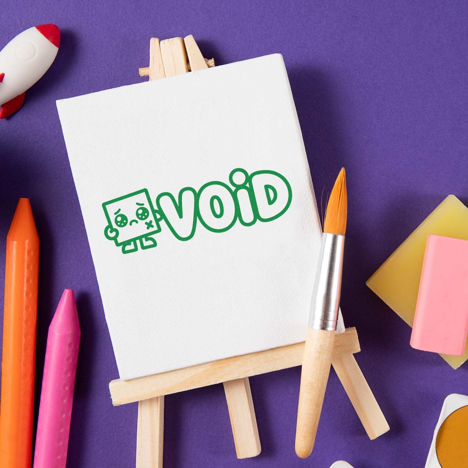 White canvas easel with green VOID stamp-and-seal logo, surrounded by colorful crayons and brushes on a purple background.