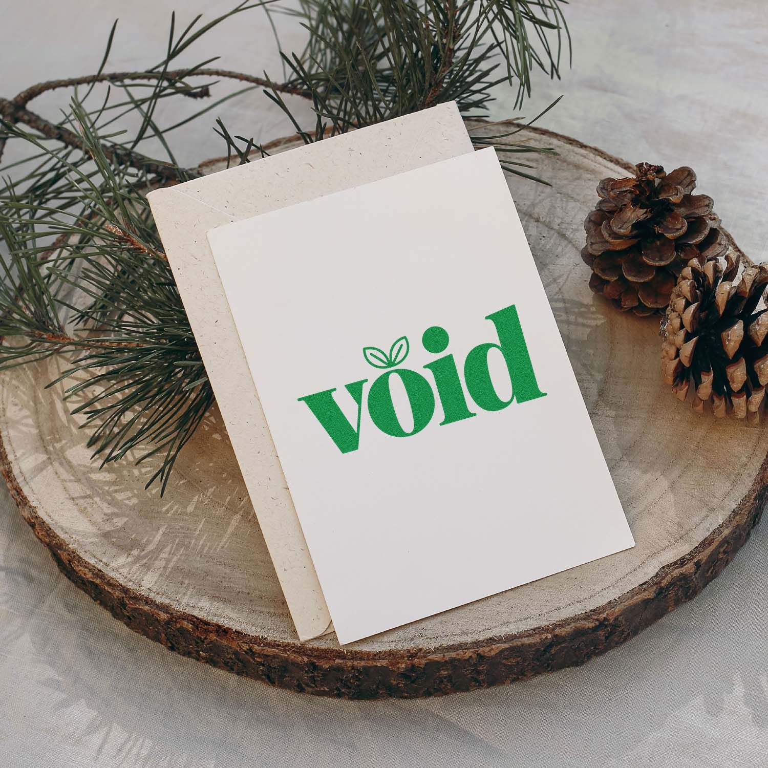White stamp and seal cards with green void logo, set on a wood slice with pine branches and pinecones.