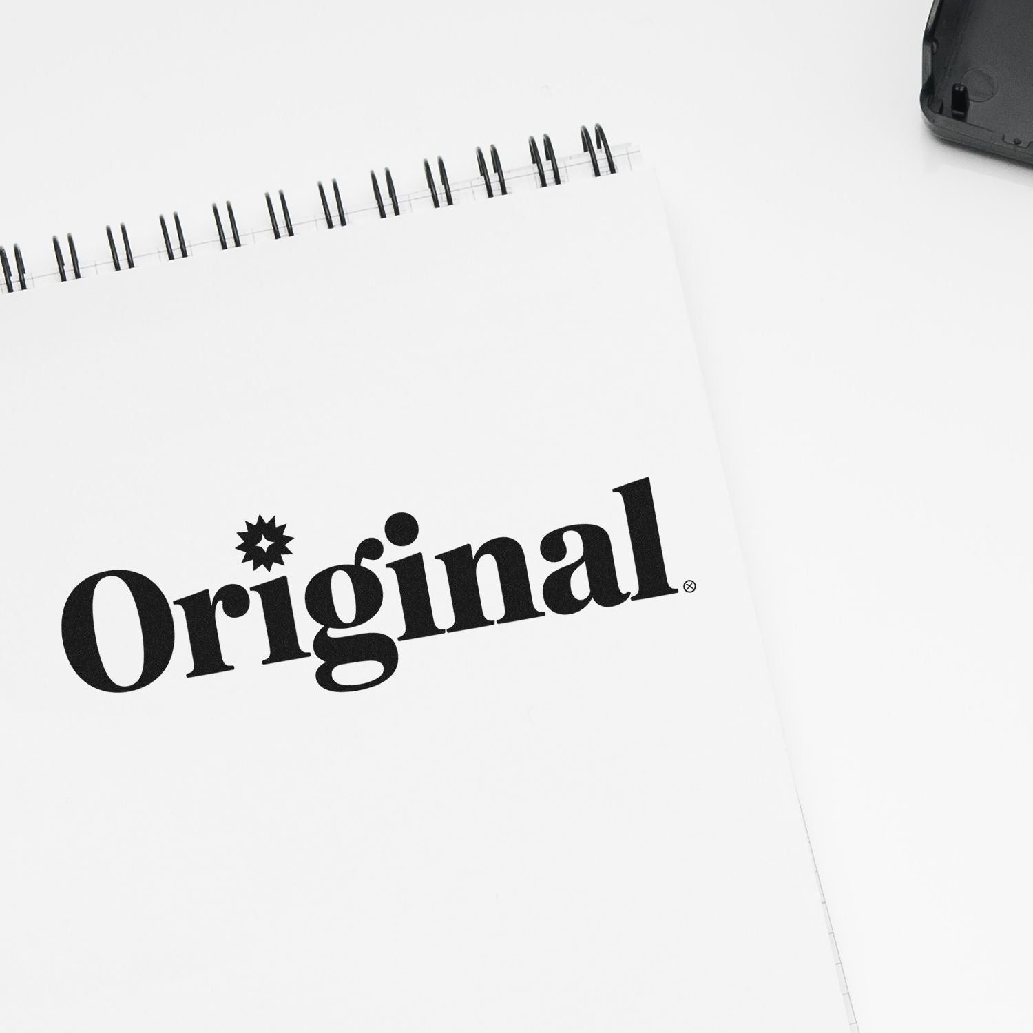 Black Original stamp and seal logo on a white spiral notebook, with a black product case in the corner.