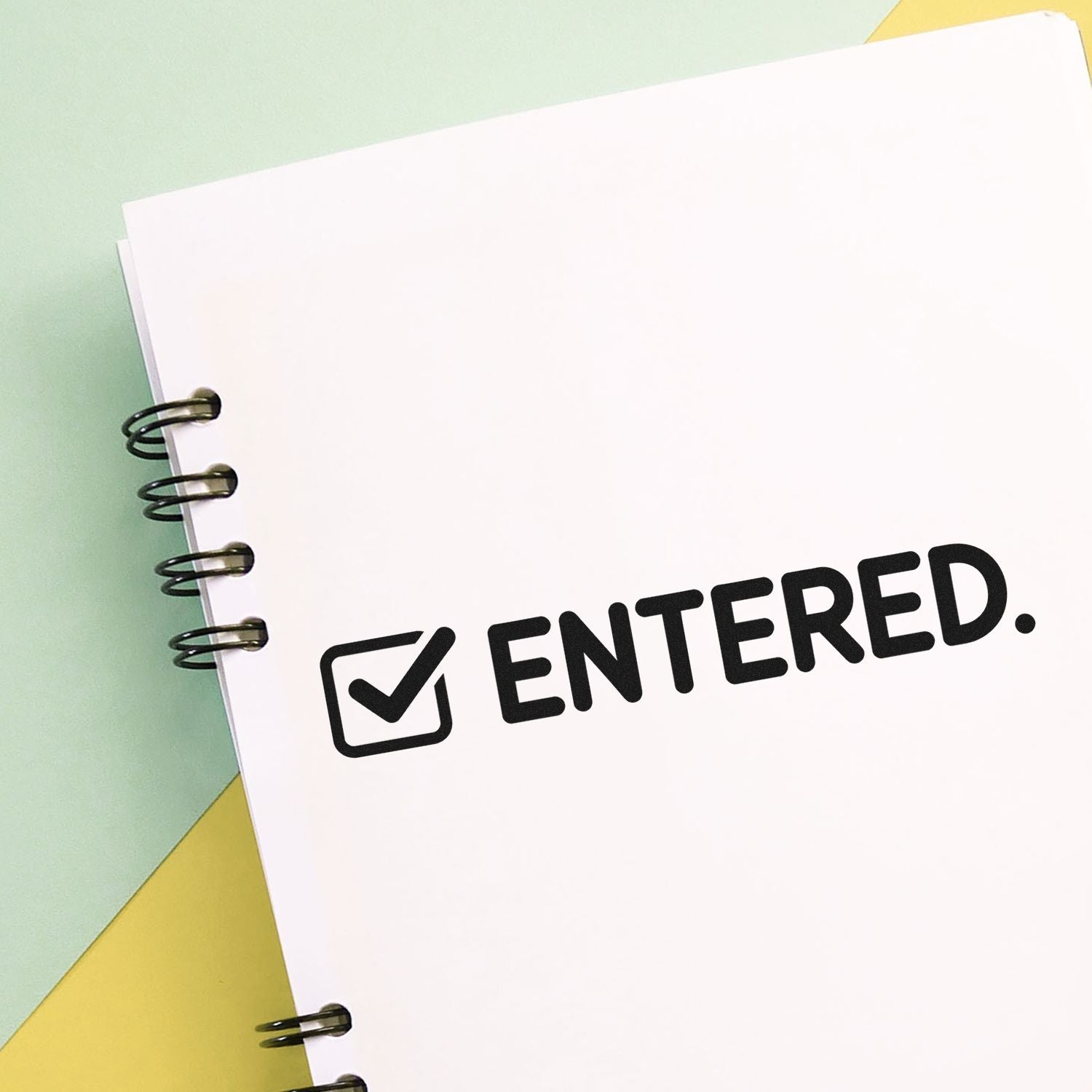 Black ENTERED self-inking stamp with checkmark on a white spiral notebook, over mint green and yellow background.