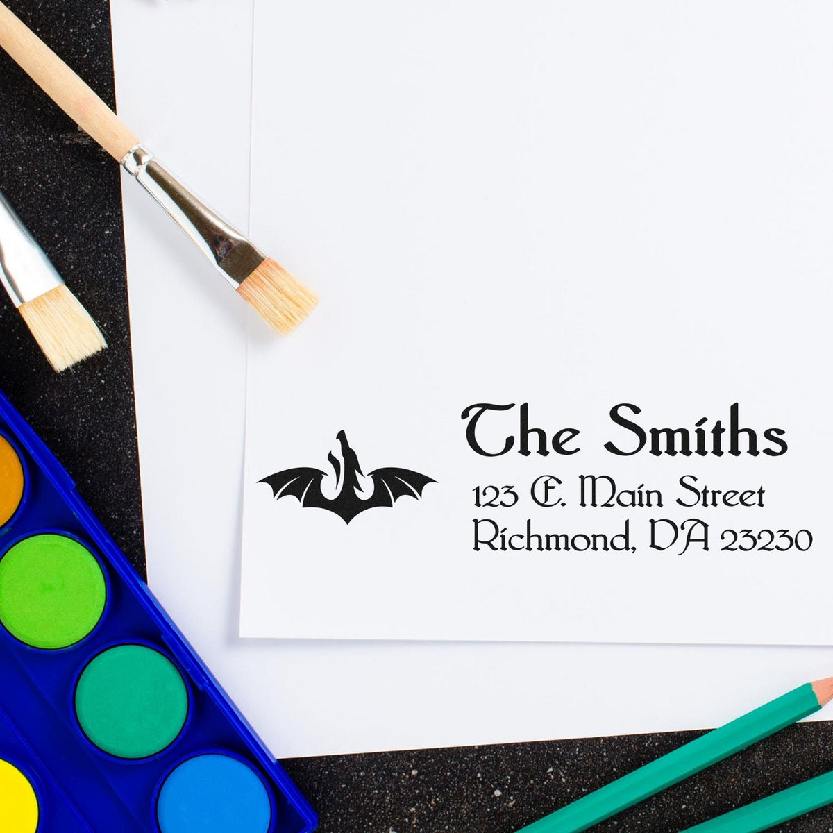 Custom stamp and wax seal for The Smiths, black bat logo on white paper with paintbrushes and bright watercolor palette.