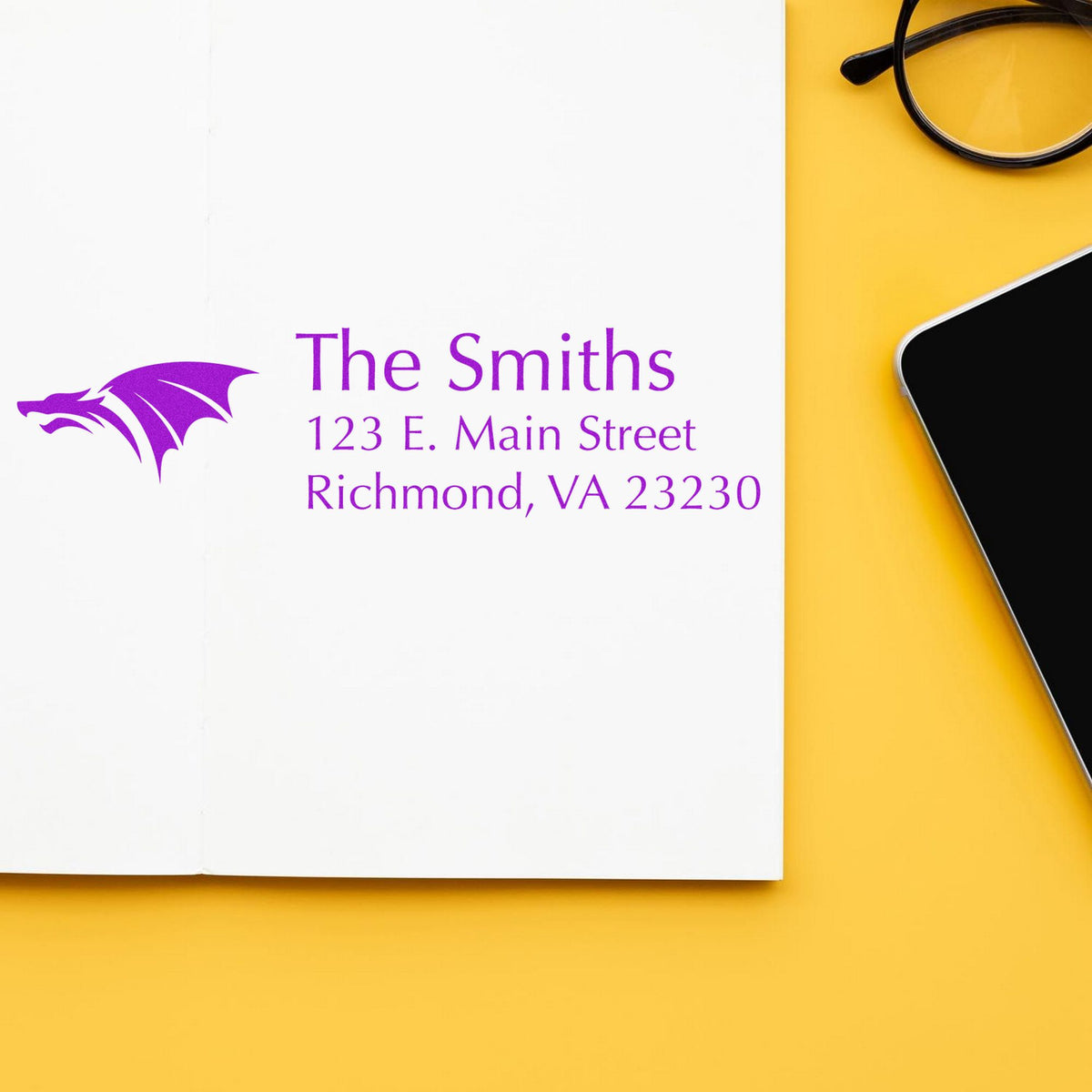 Custom purple address stamp and seal for The Smiths, with dragon logo, on white paper beside glasses and a phone on yellow background.