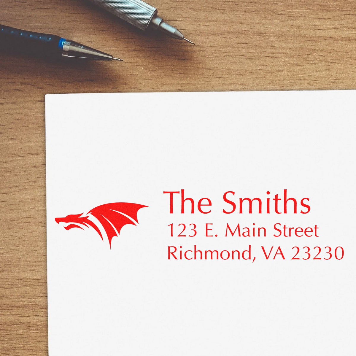 Red custom address stamp and seal with dragon logo on white paper, beside two silver pens on a wood desk.