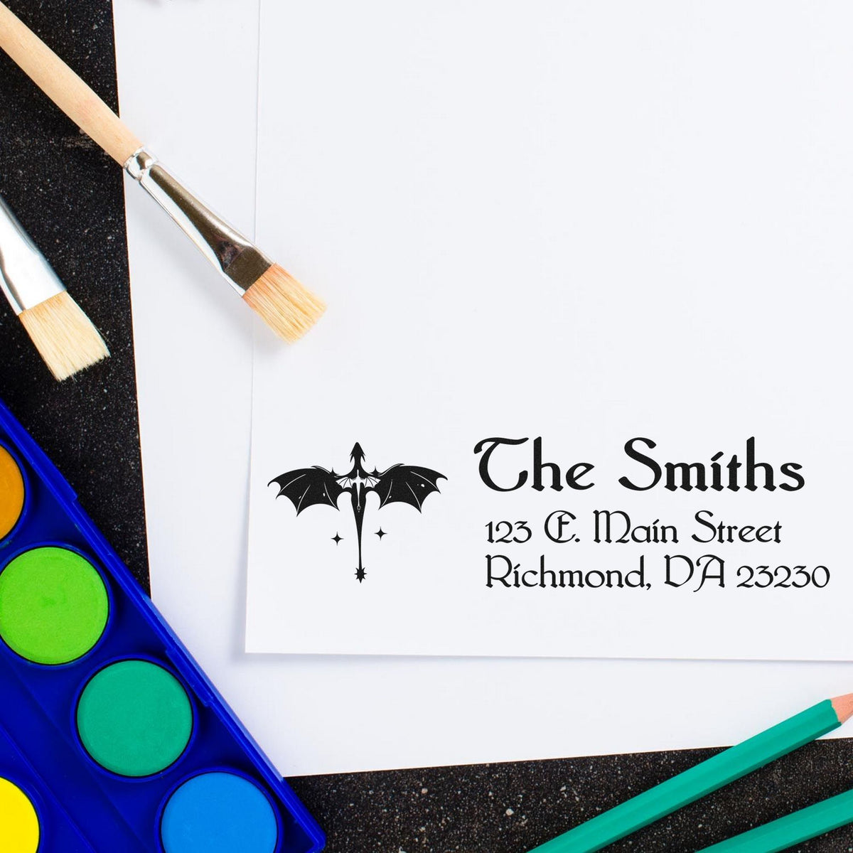 Custom black stamp and seal with bat logo and gothic text on white paper, surrounded by paintbrushes and bright watercolor paints.