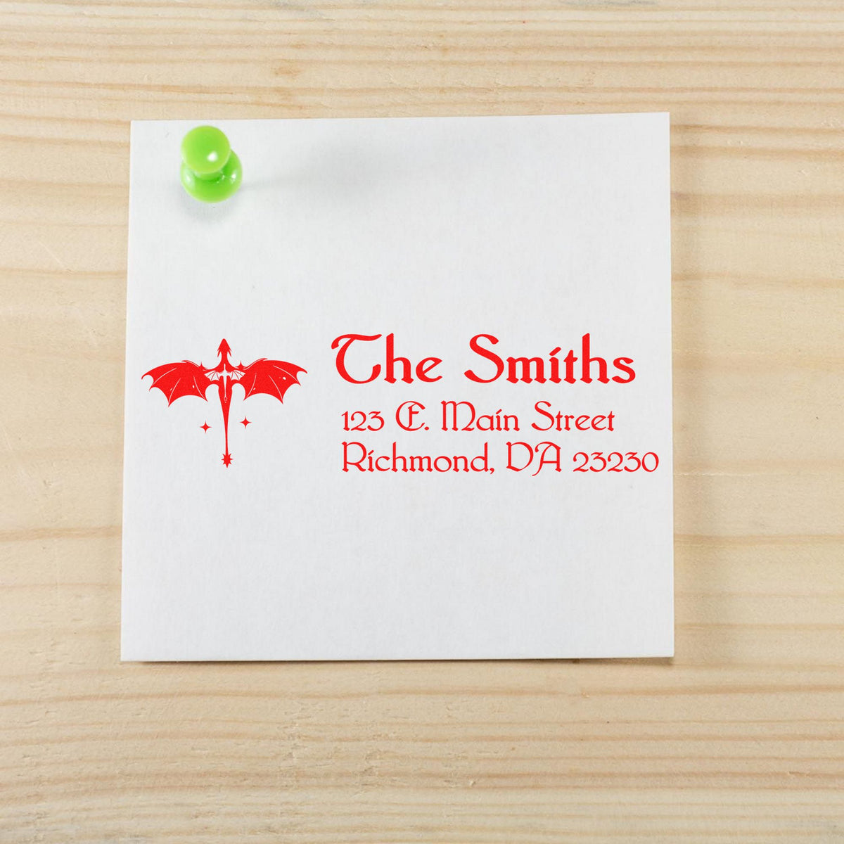 White paper with red custom return address stamp, bat emblem, and green pushpin on a light wood background.