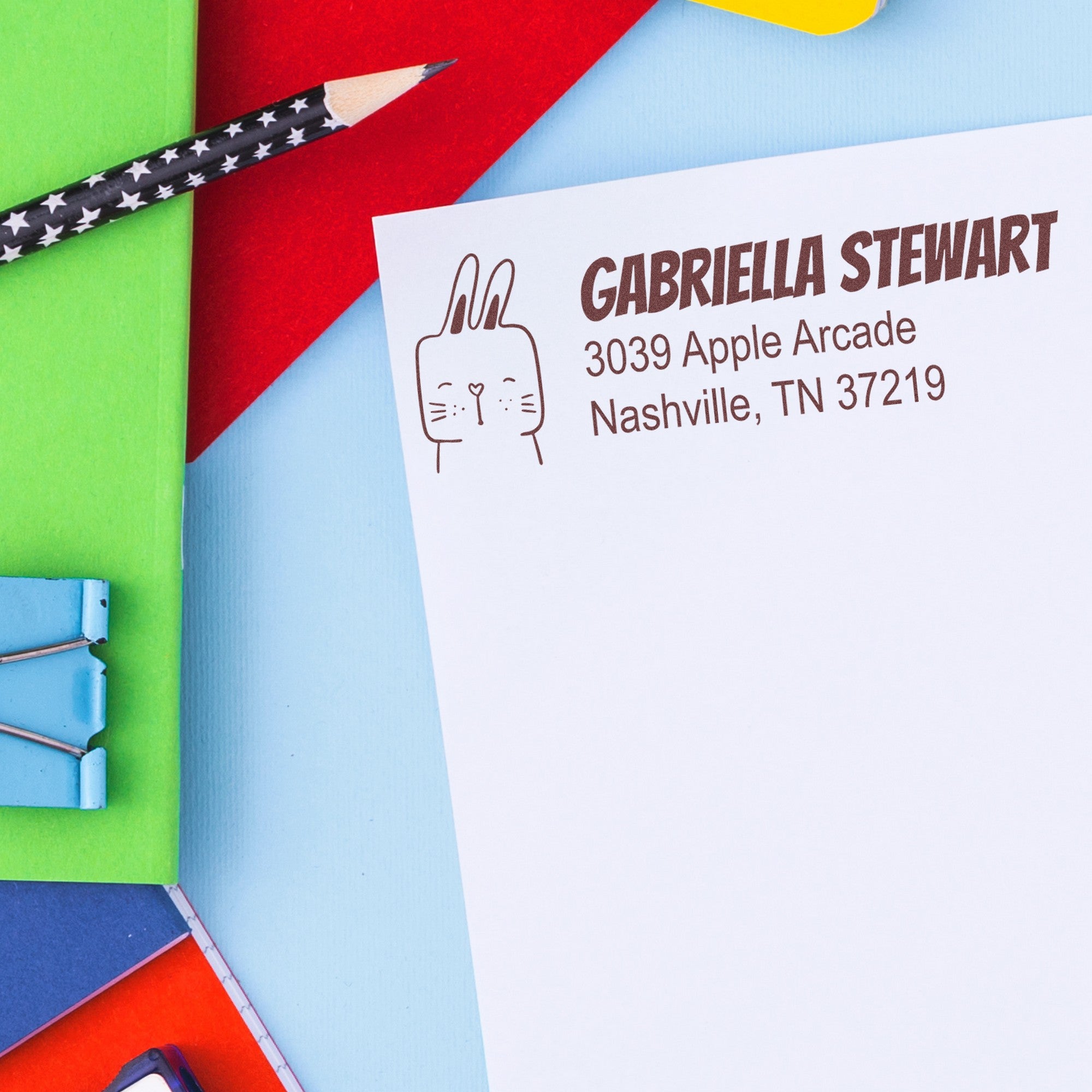 Fuzzy Rabbit Custom Mail Address Self-Inking Stamp on a letter with "GABRIELLA STEWART, 3039 Apple Arcade, Nashville, TN 37219." The Customized Home Stamper design features a cute rabbit illustration. Surrounding items include a star-patterned pencil and colorful paper.