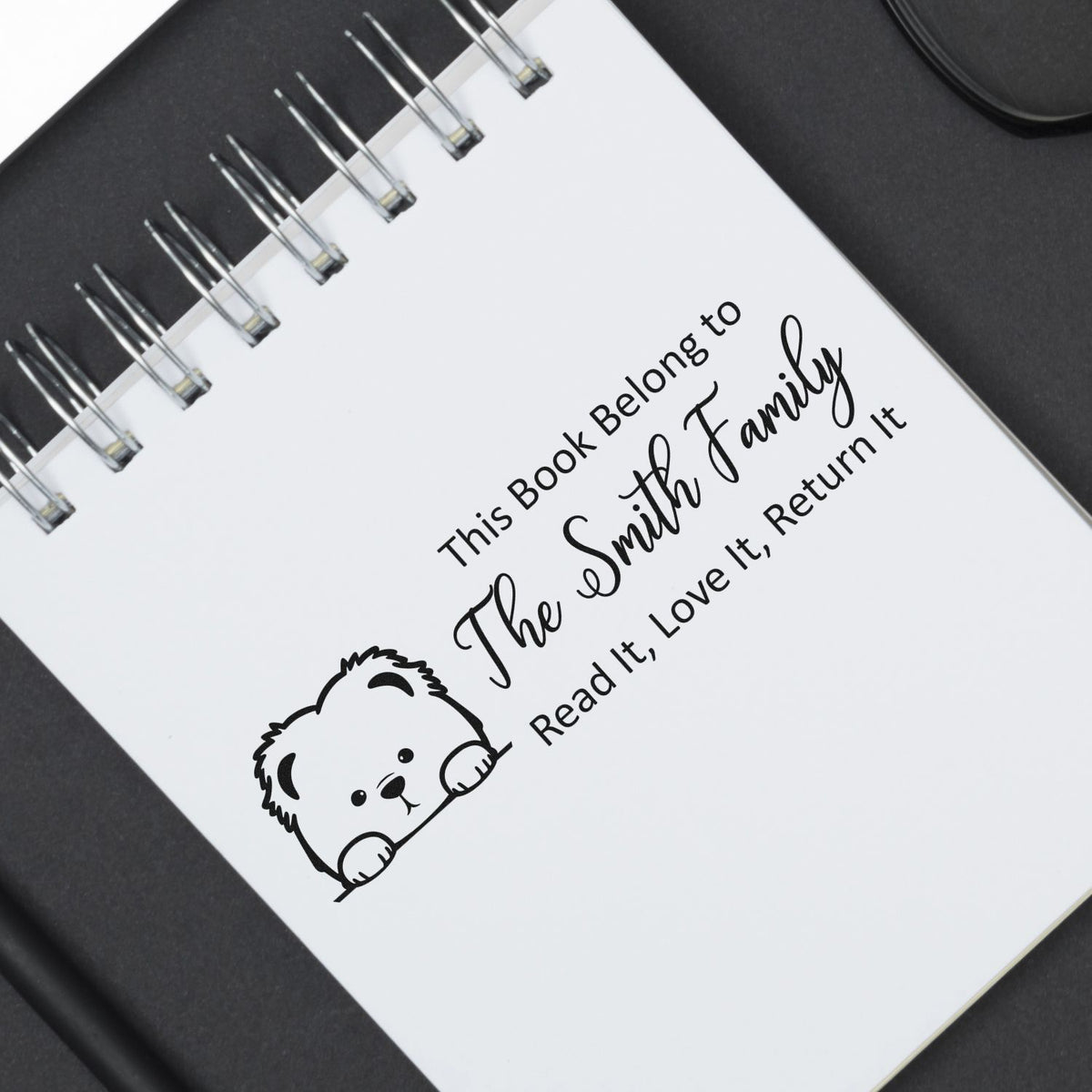 Black book stamp seal on a white spiral notebook, reading This Book Belongs to The Smith Family with a cute bear.
