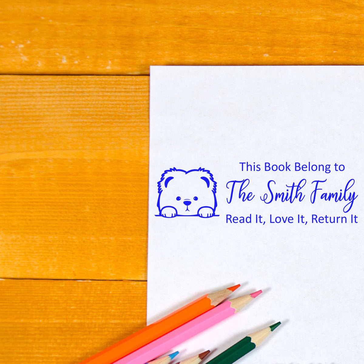 Blue personalized book stamp with bear icon and The Smith Family text on white paper, beside colorful pencils on a wood desk.