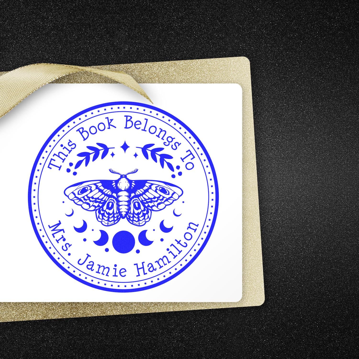 Blue custom bookplate stamp seal with moth, moon phases, and This Book Belongs To Mrs. Jamie Hamilton on a white card.