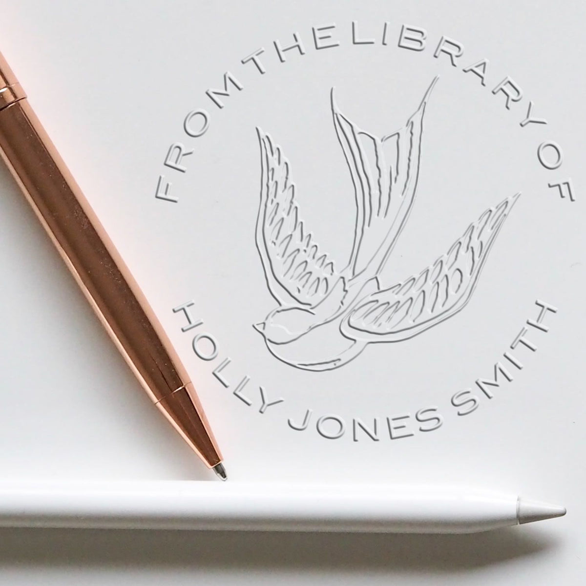 Embossed white stamp seal with flying bird logo and Holly Jones Smith text, paired with a rose gold pen.