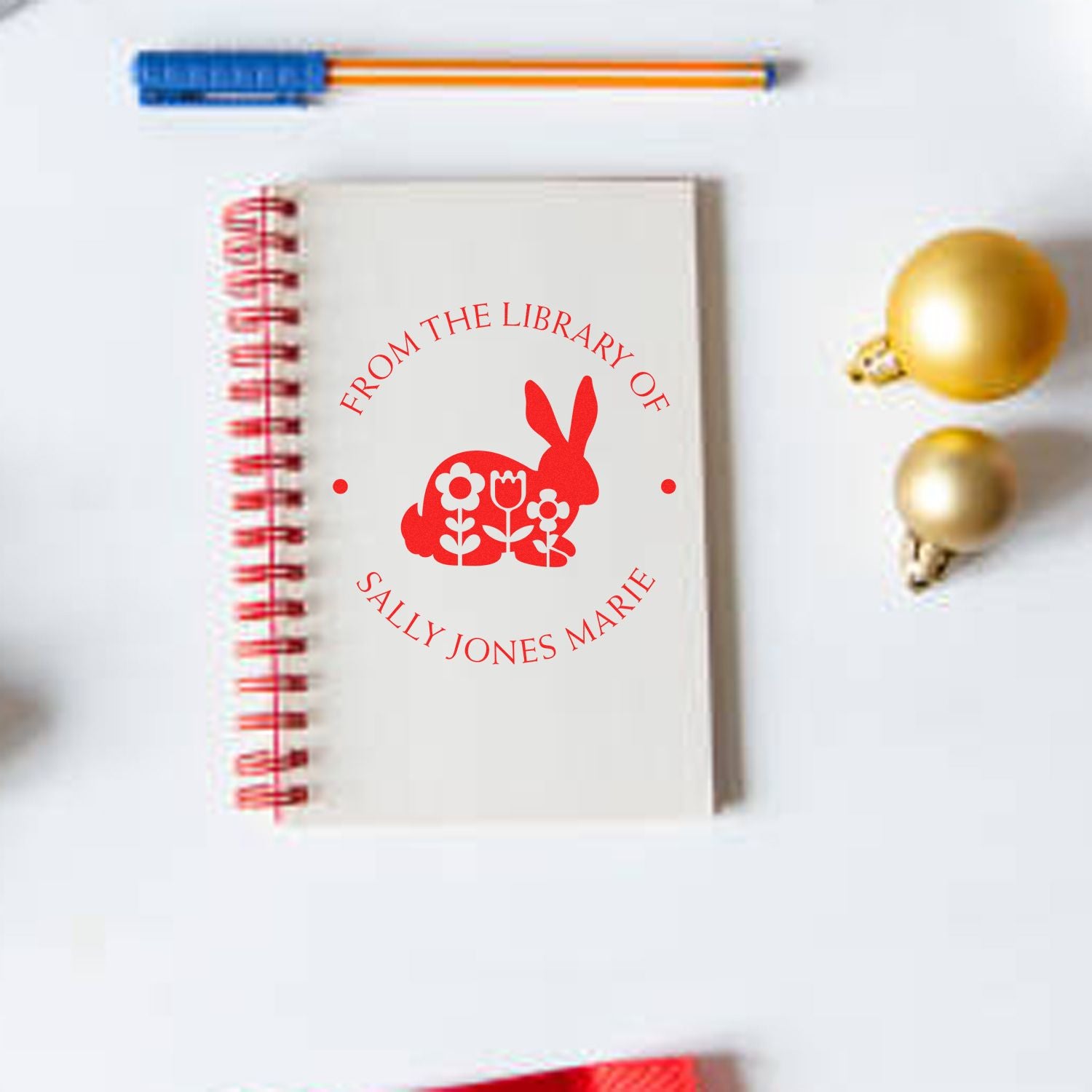 Custom red library stamp on cream notebook with rabbit and flowers; blue pen and gold ornaments nearby.
