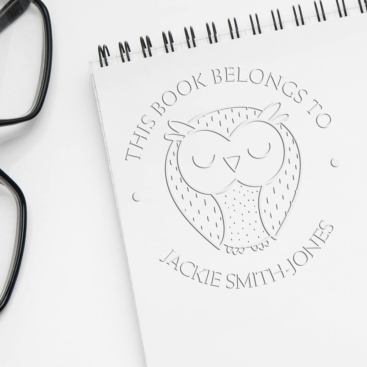 White custom book embosser seal with owl design and This Book Belongs to Jackie Smith-Jones text, beside black eyeglasses.