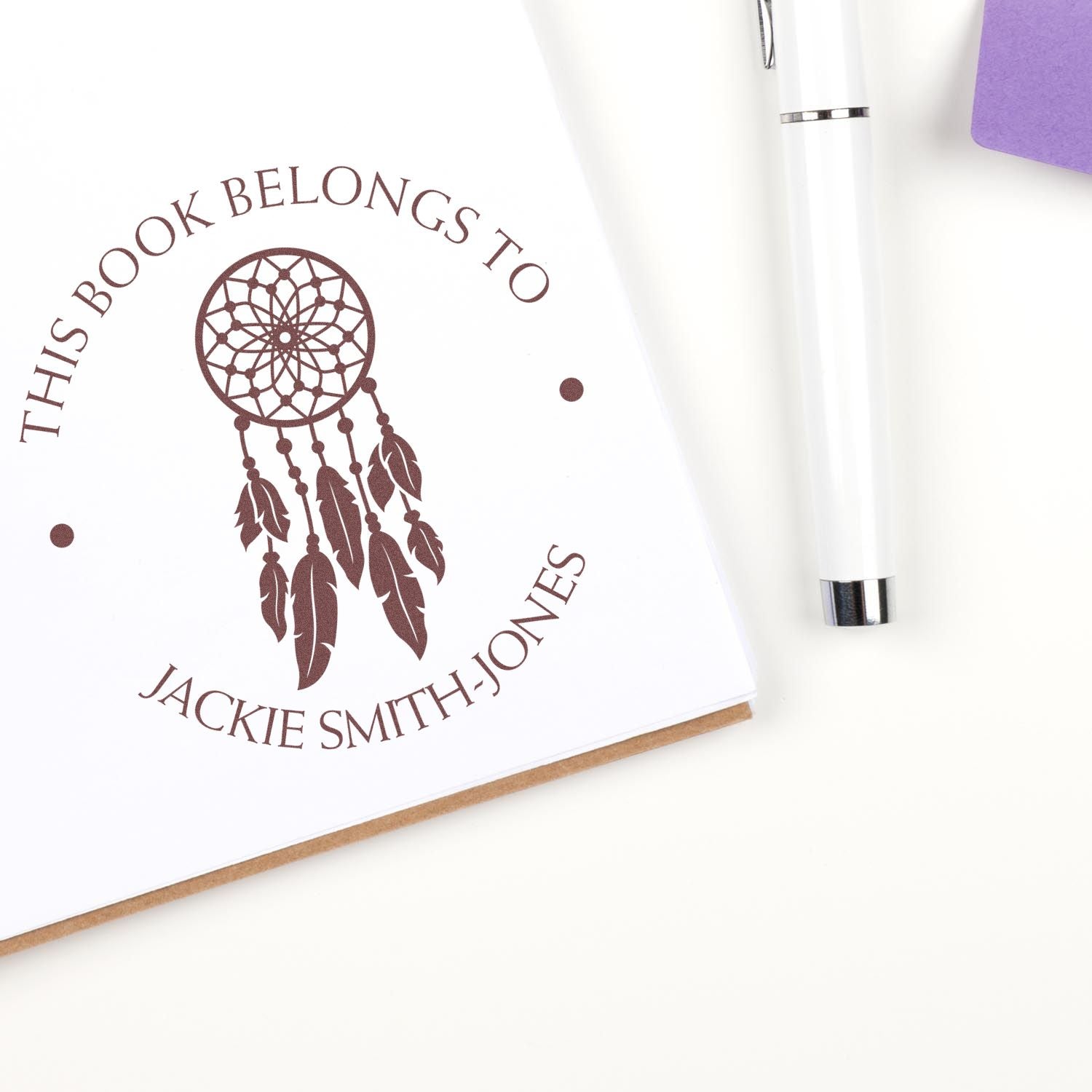 Brown custom book-belongs-to stamp seal with dreamcatcher on white card, beside a white silver pen and purple envelope.